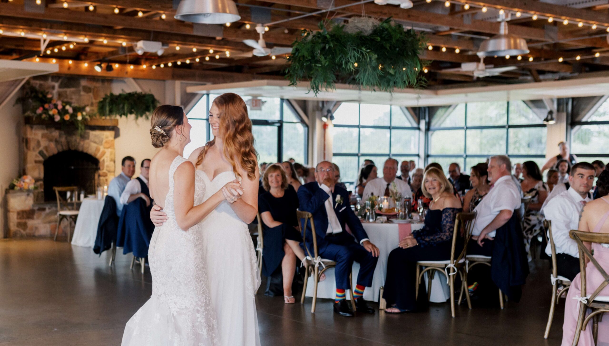 Two brides share a dance at a wedding reception in a decorated venue with guests seated at tables in the background.