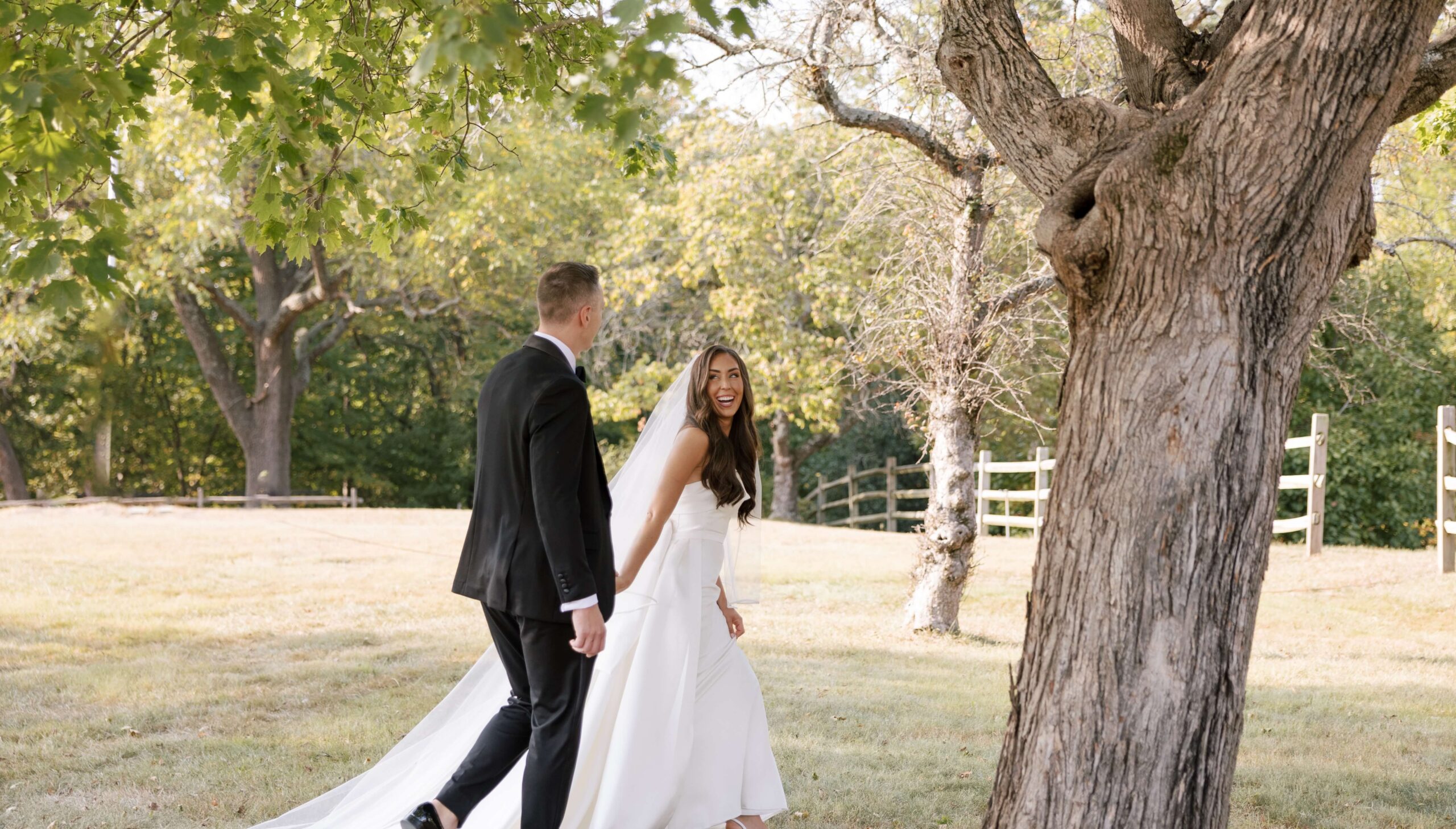 A bride and groom walk hand-in-hand in a grassy area with trees and a fence in the background. The bride is smiling and wearing a white dress; the groom is in a black suit.