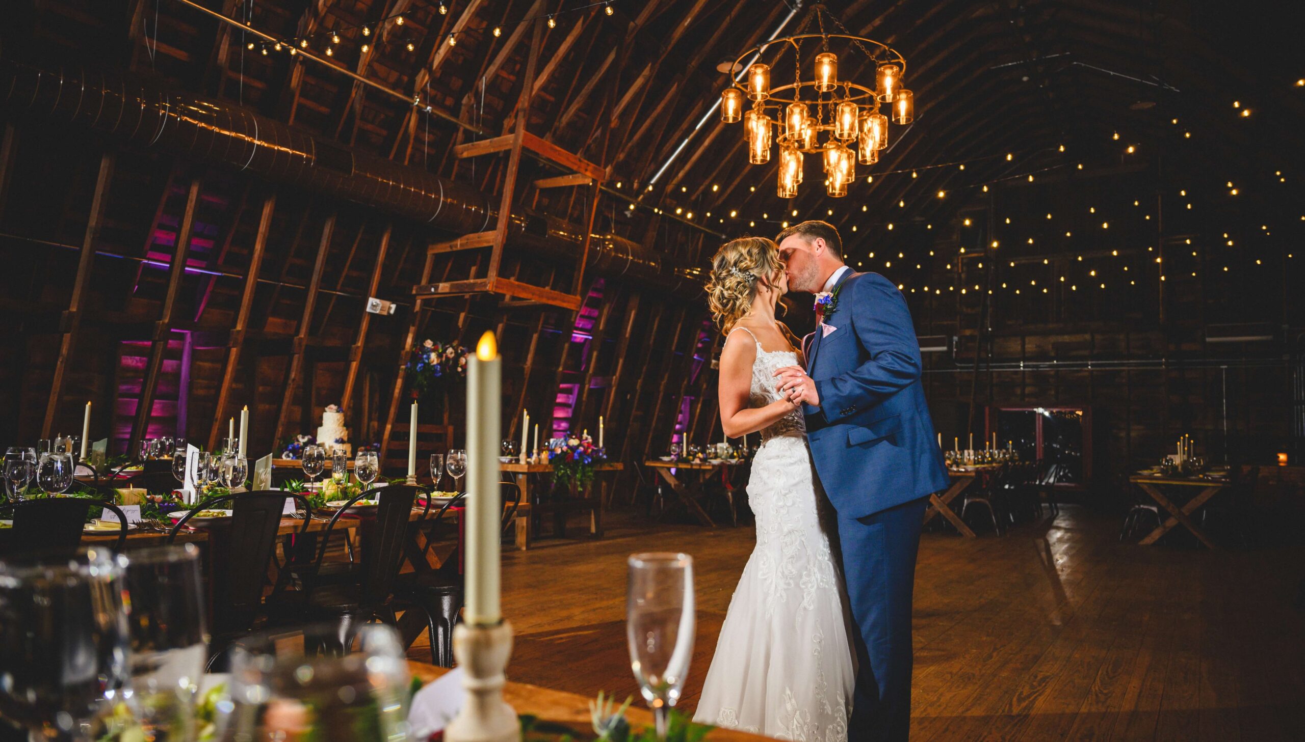 A bride and groom share their first dance in a warmly lit barn, surrounded by tables set for a wedding reception, with candles and string lights overhead.