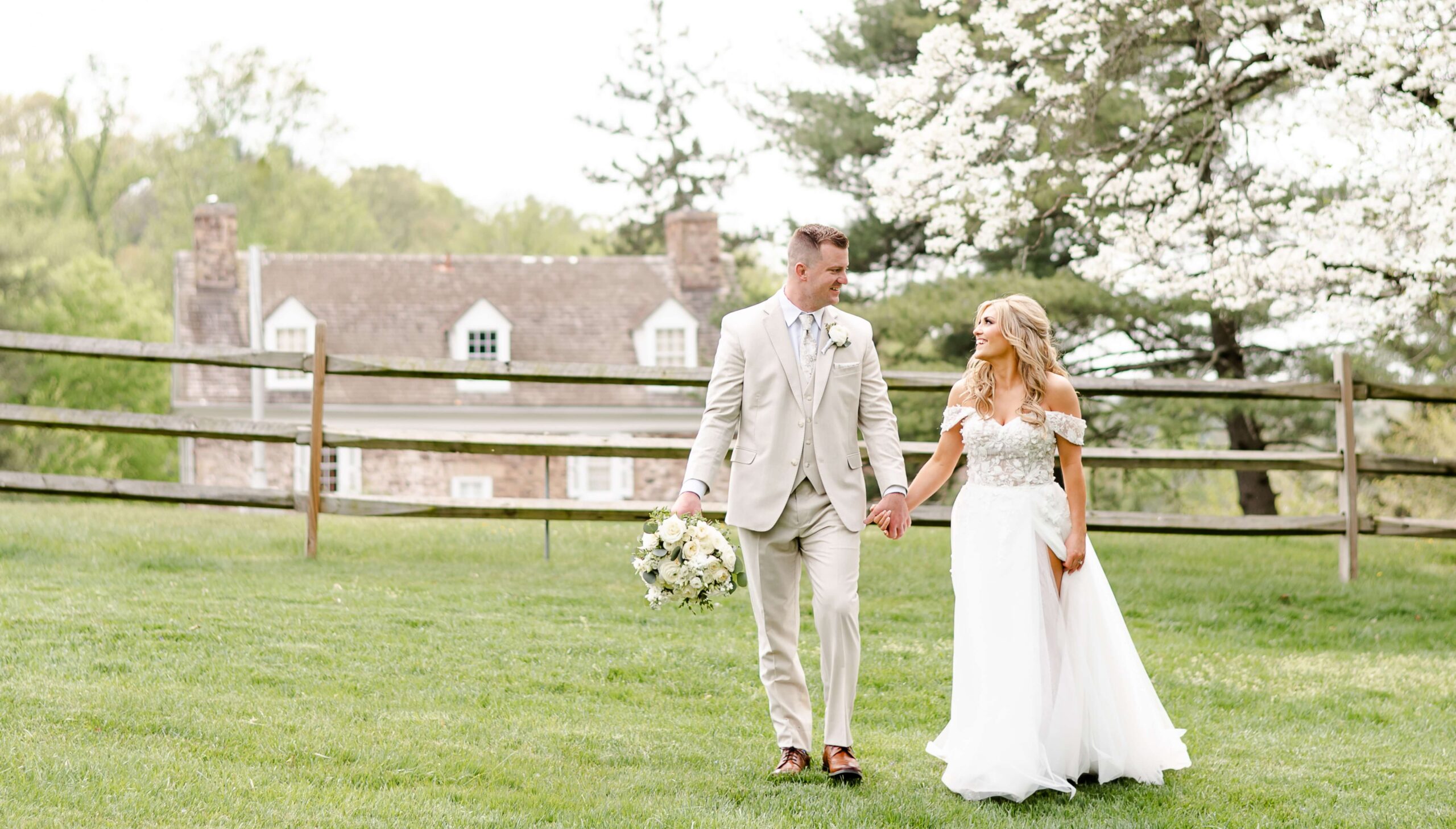 A bride and groom walk hand in hand on grass, with a wooden fence and a large house in the background, surrounded by trees. The groom holds a bouquet.