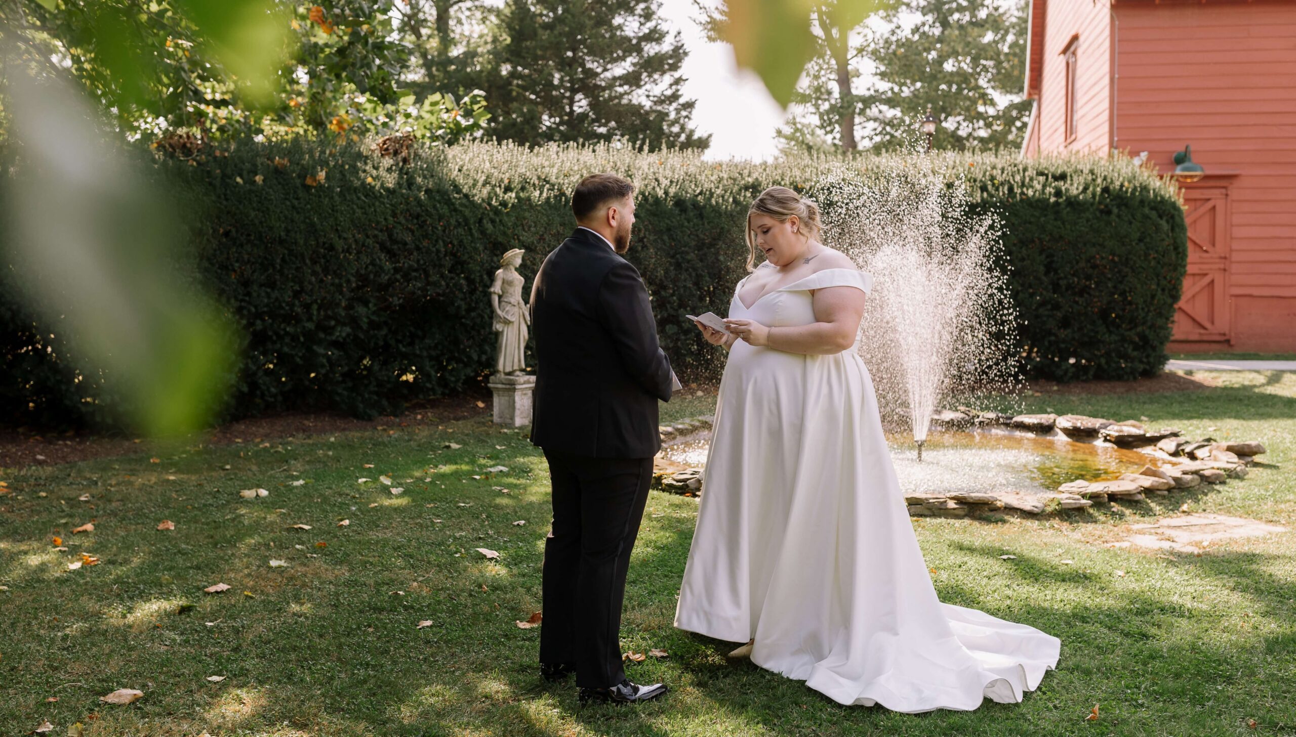 A bride and groom exchange vows in a garden setting with a fountain and greenery in the background.