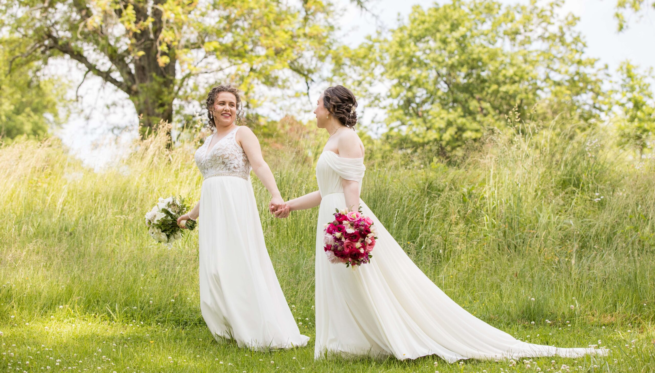 Two people in white bridal gowns holding hands, walking on grass in a park. One is holding a bouquet of white flowers and the other a bouquet of red flowers. Trees are in the background.