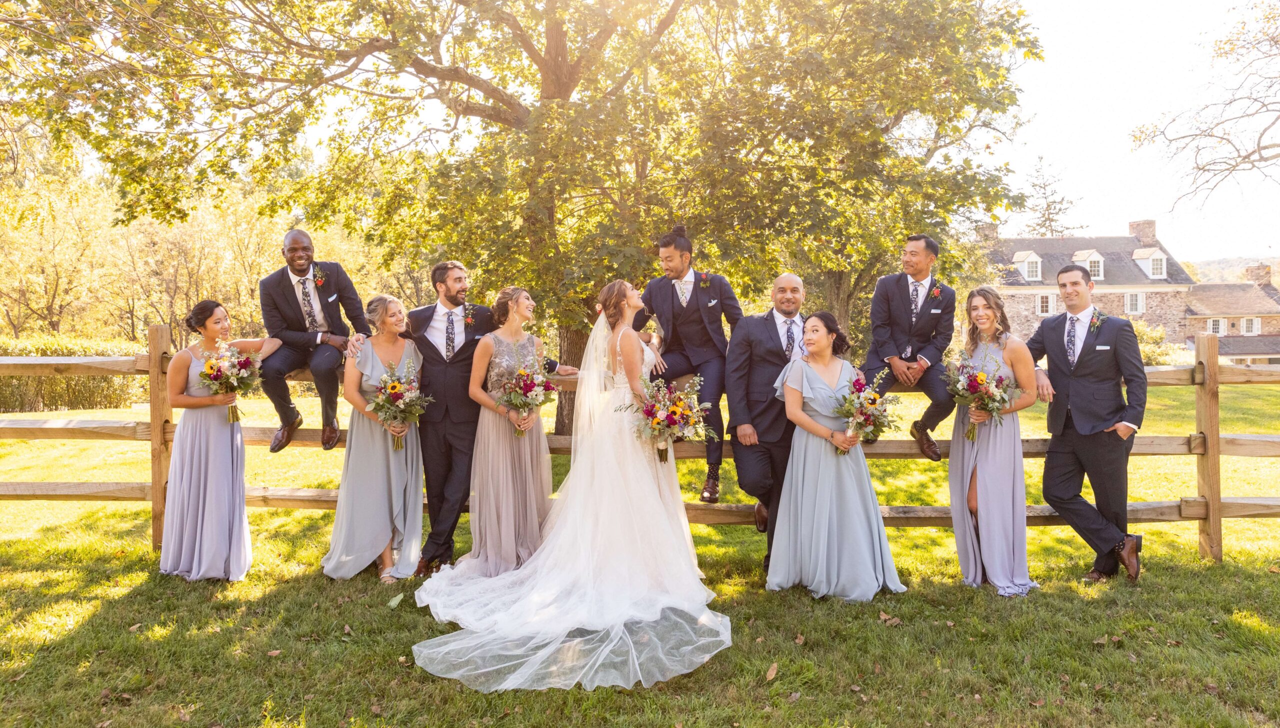 Wedding party poses outdoors by a wooden fence, dressed in formal attire. The bride and groom stand together, surrounded by bridesmaids holding bouquets and groomsmen wearing suits.