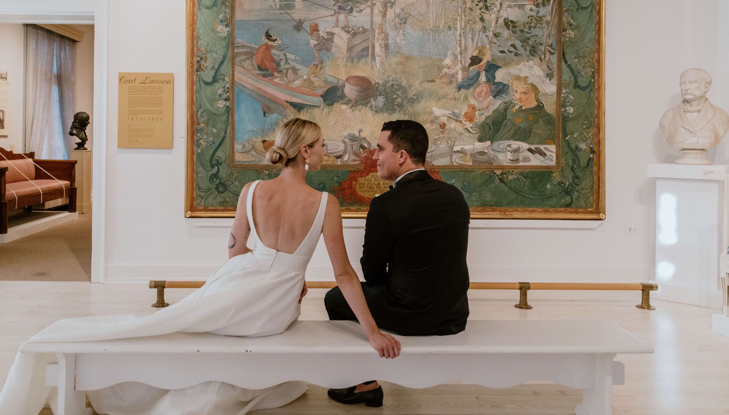 A couple sits on a bench, facing a large tapestry in a gallery. The woman is in a white dress; the man in a black outfit. Bust sculptures are visible in the background.