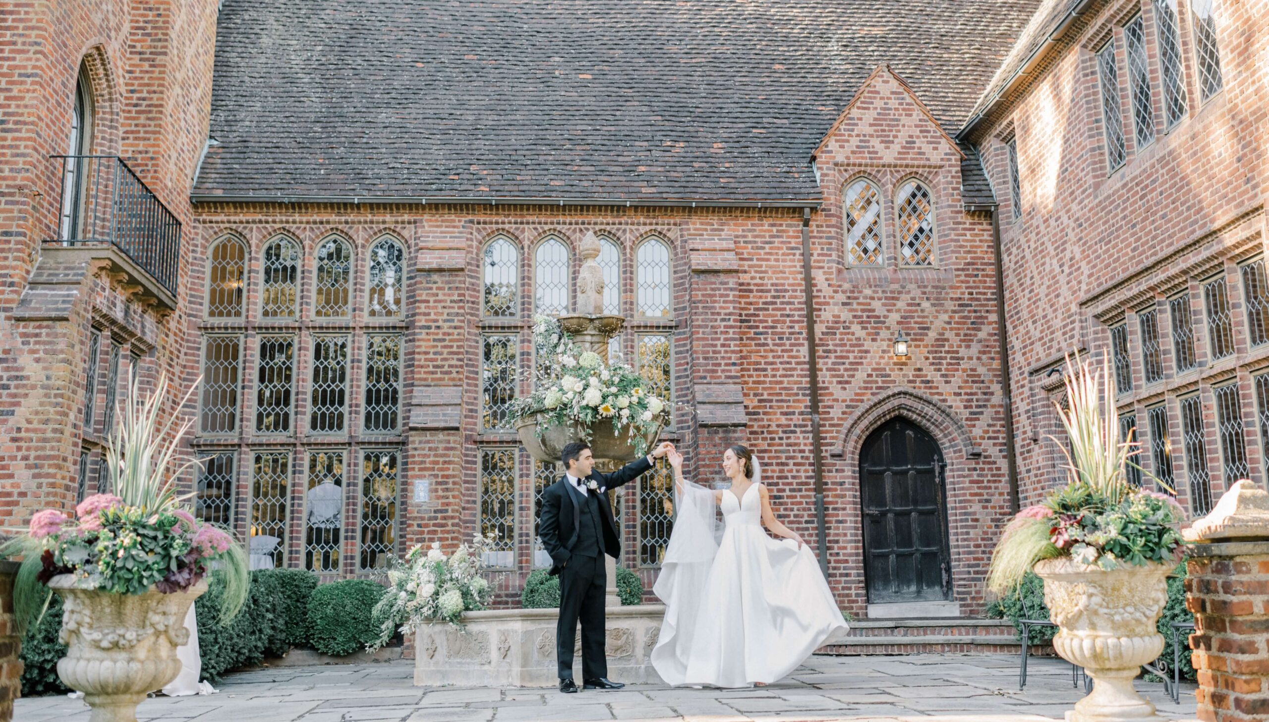 A bride and groom dance in a courtyard in front of a historic brick building with large windows and a floral fountain.