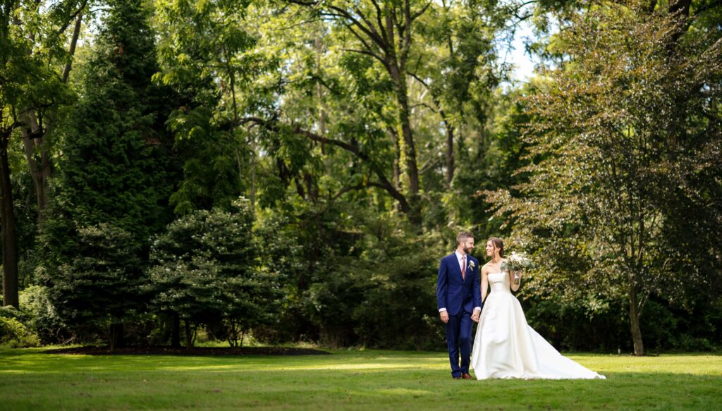 A bride and groom walk together on a grassy area in a park, surrounded by trees.