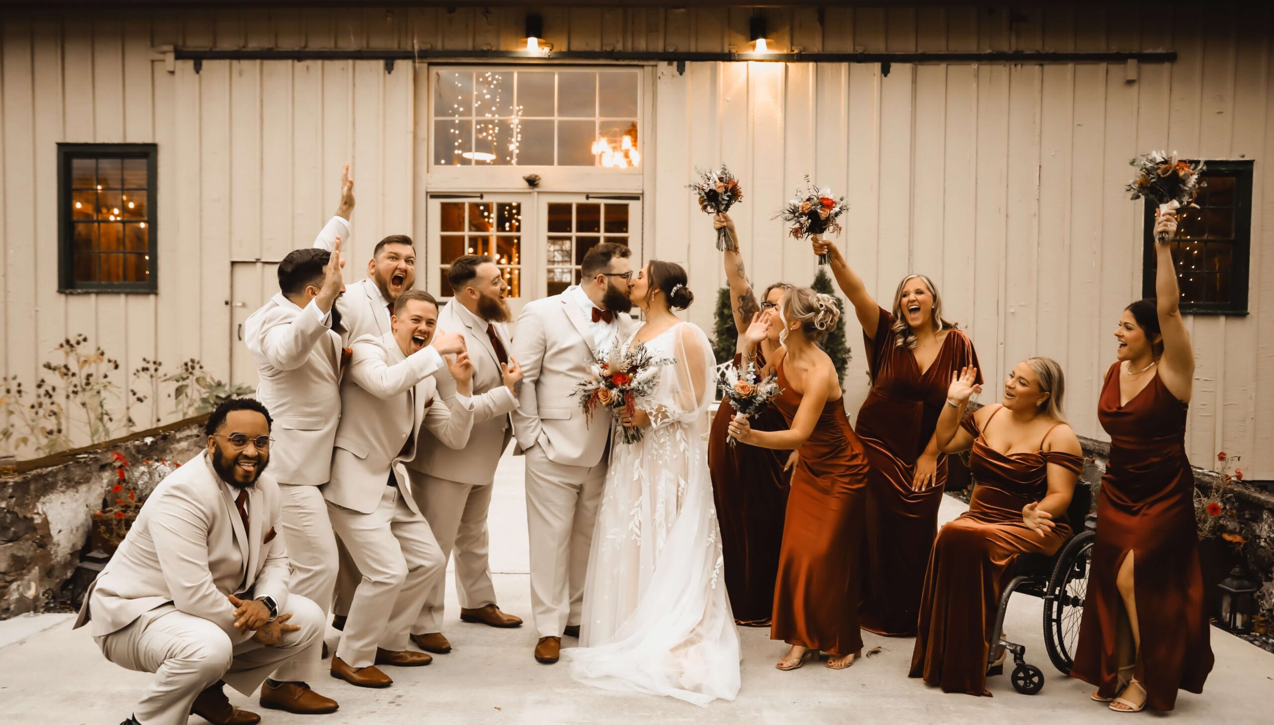 A wedding party poses joyfully outside a rustic building. The bride and groom kiss in the center, surrounded by bridesmaids in brown dresses and groomsmen in beige suits.