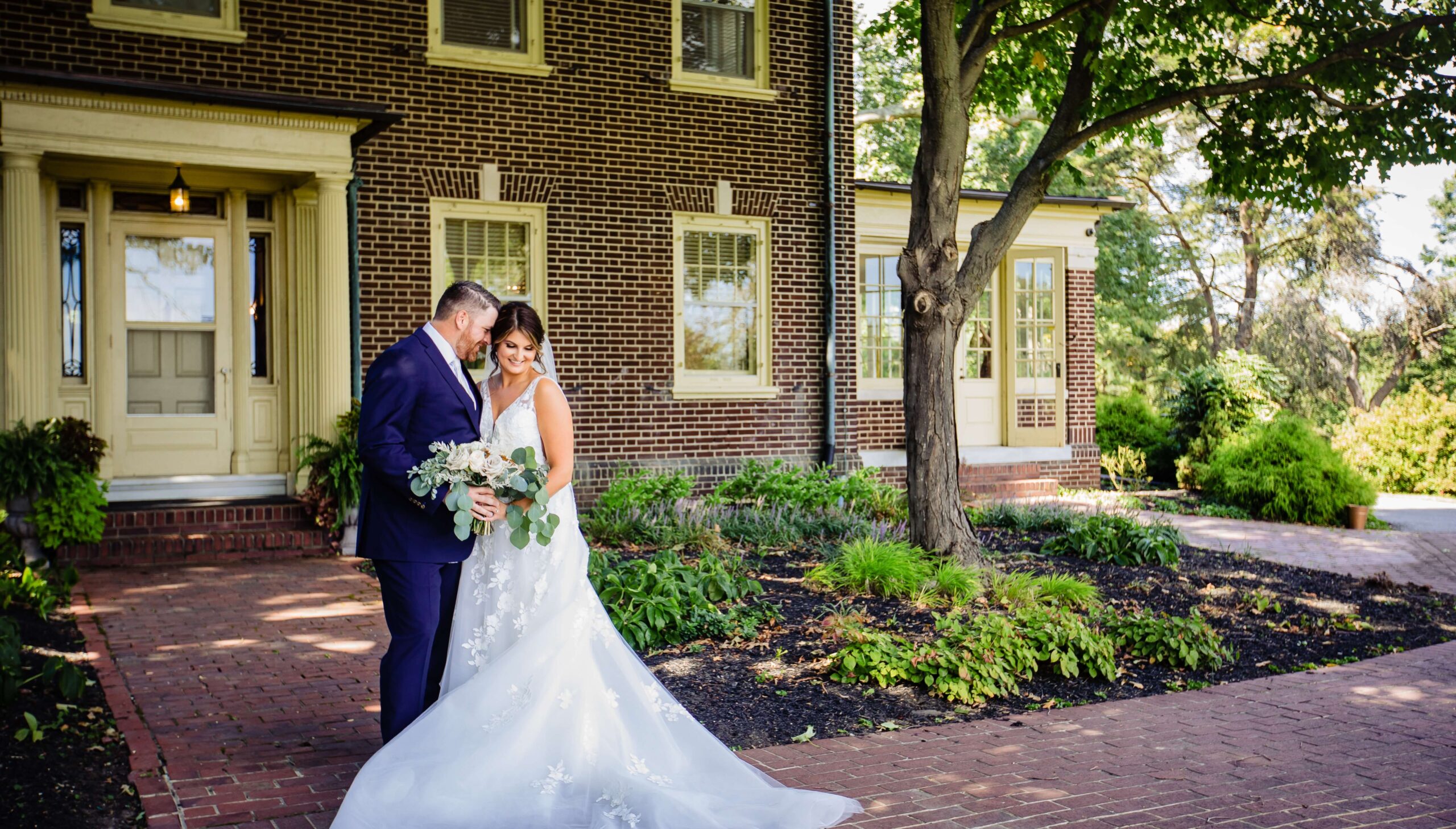 A bride and groom stand in front of a brick house with a large tree, posing for a photo on a sunny day. The bride holds a bouquet and wears a white gown.
