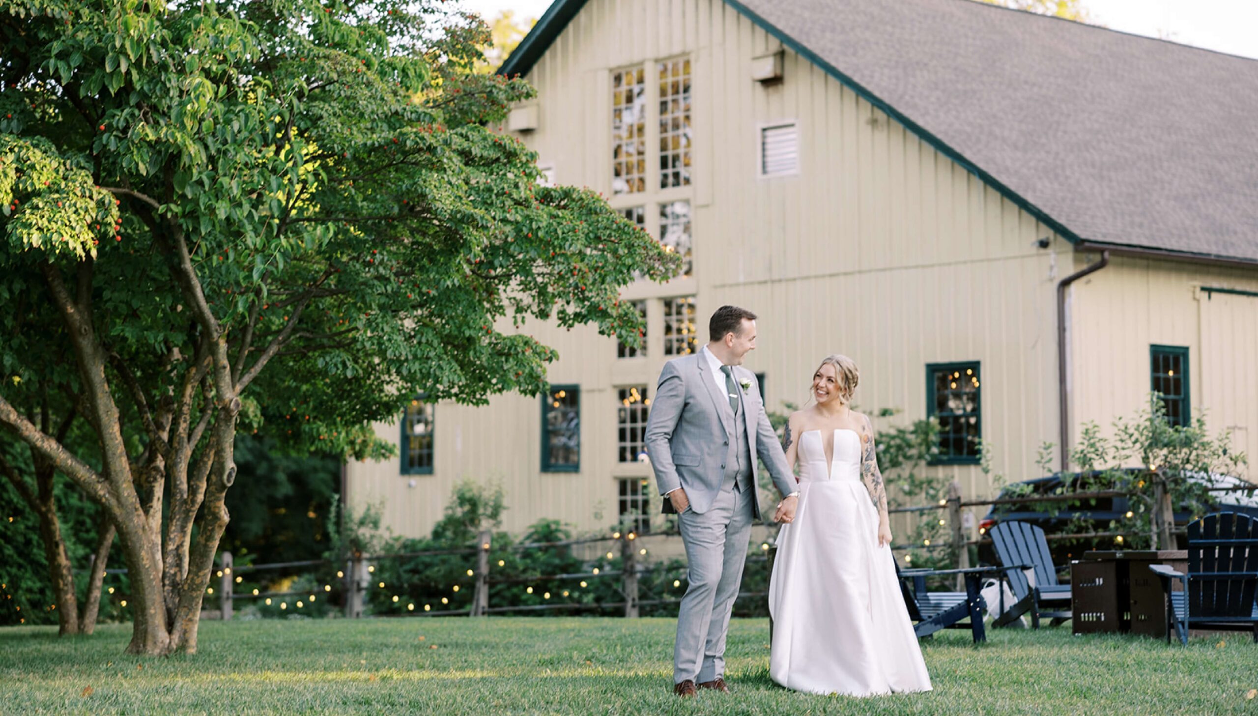 A couple in wedding attire holding hands and walking on grass, with a light-colored barn and trees in the background.