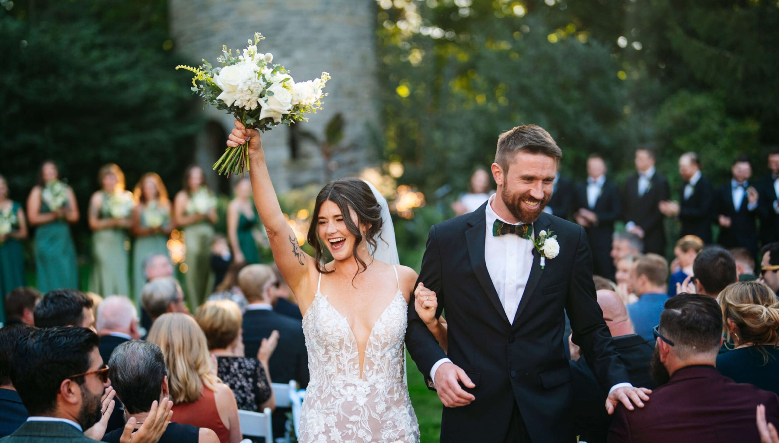 A newlywed couple walks down the aisle outdoors, with the bride raising a bouquet. Guests are seated and clapping, while the bridal party stands in the background.