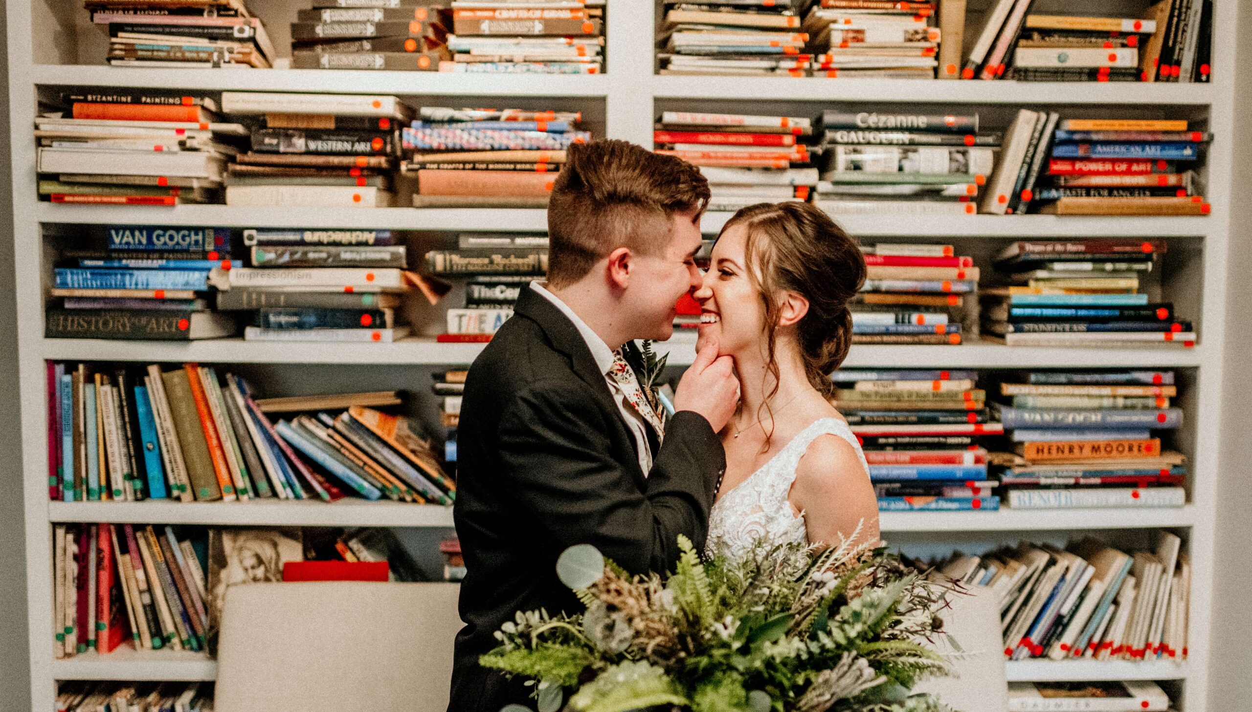 A couple in formal attire stands close, smiling at each other in front of a bookshelf filled with various books. A bouquet of flowers is in the foreground.