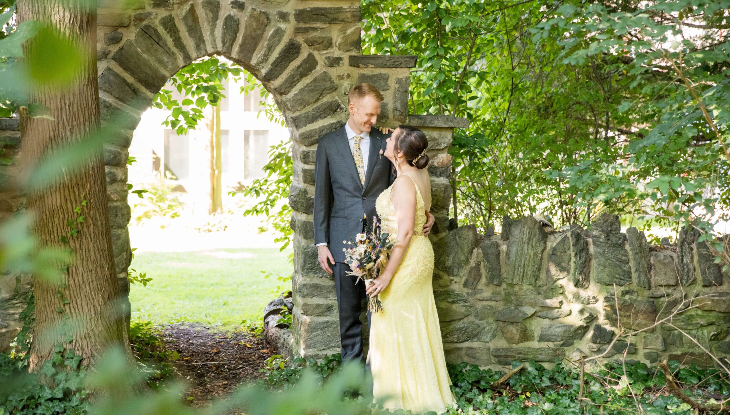 A couple stands together near a stone archway in a lush garden. The person on the left wears a suit, and the person on the right wears a yellow dress, holding a bouquet. They are smiling at each other.