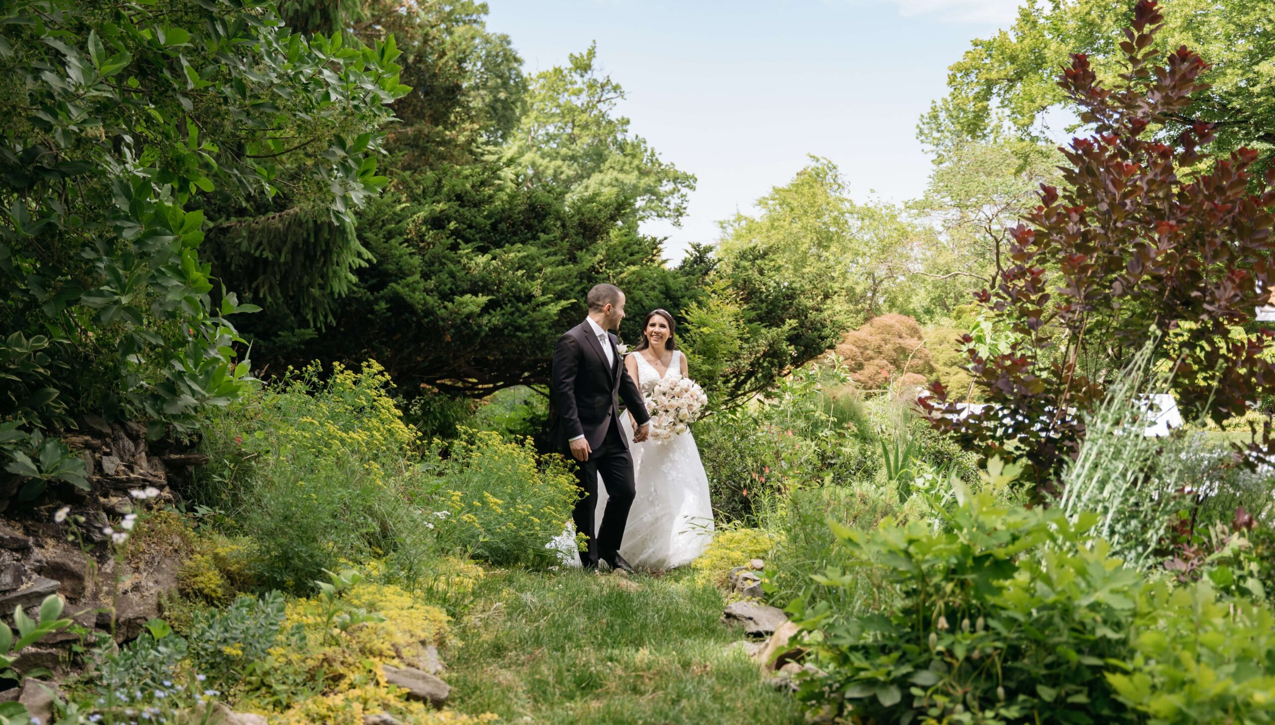 Bride and groom walking through a lush garden with greenery and flowers on both sides, under a clear blue sky.