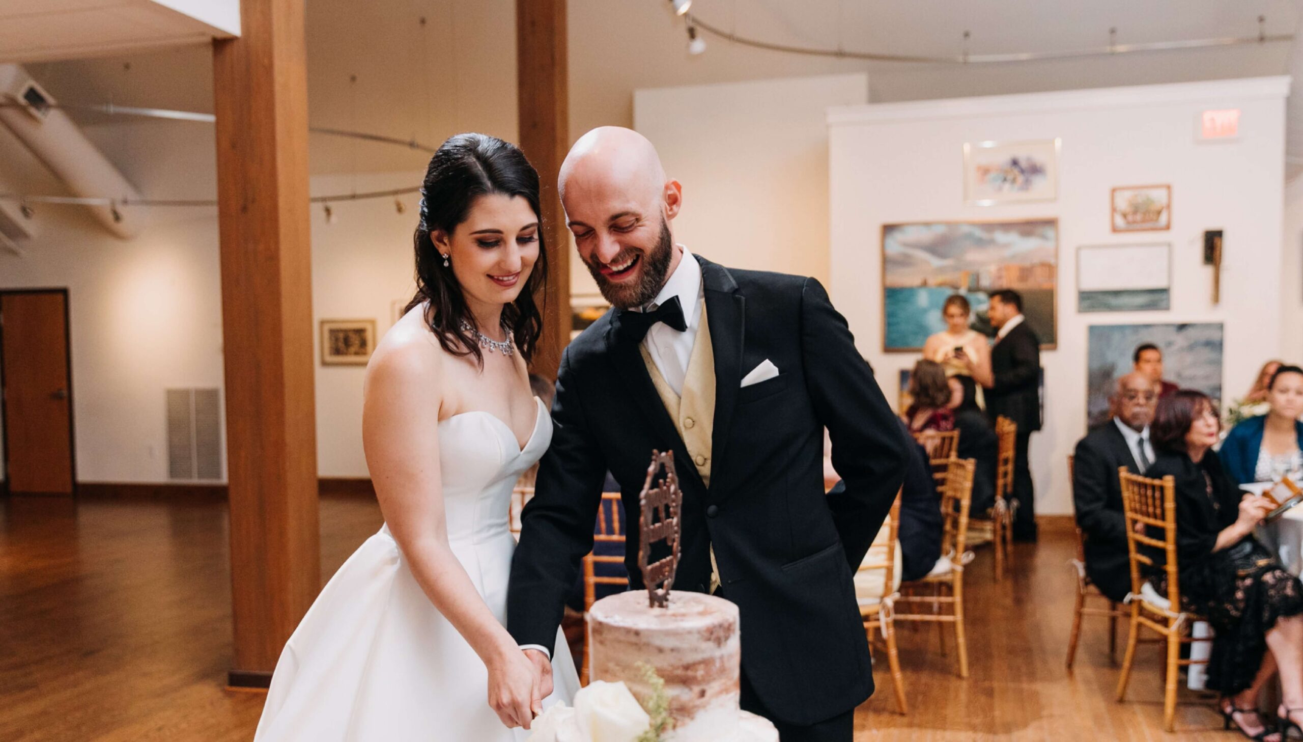 A bride and groom cut a wedding cake together in a decorated venue with guests seated in the background.