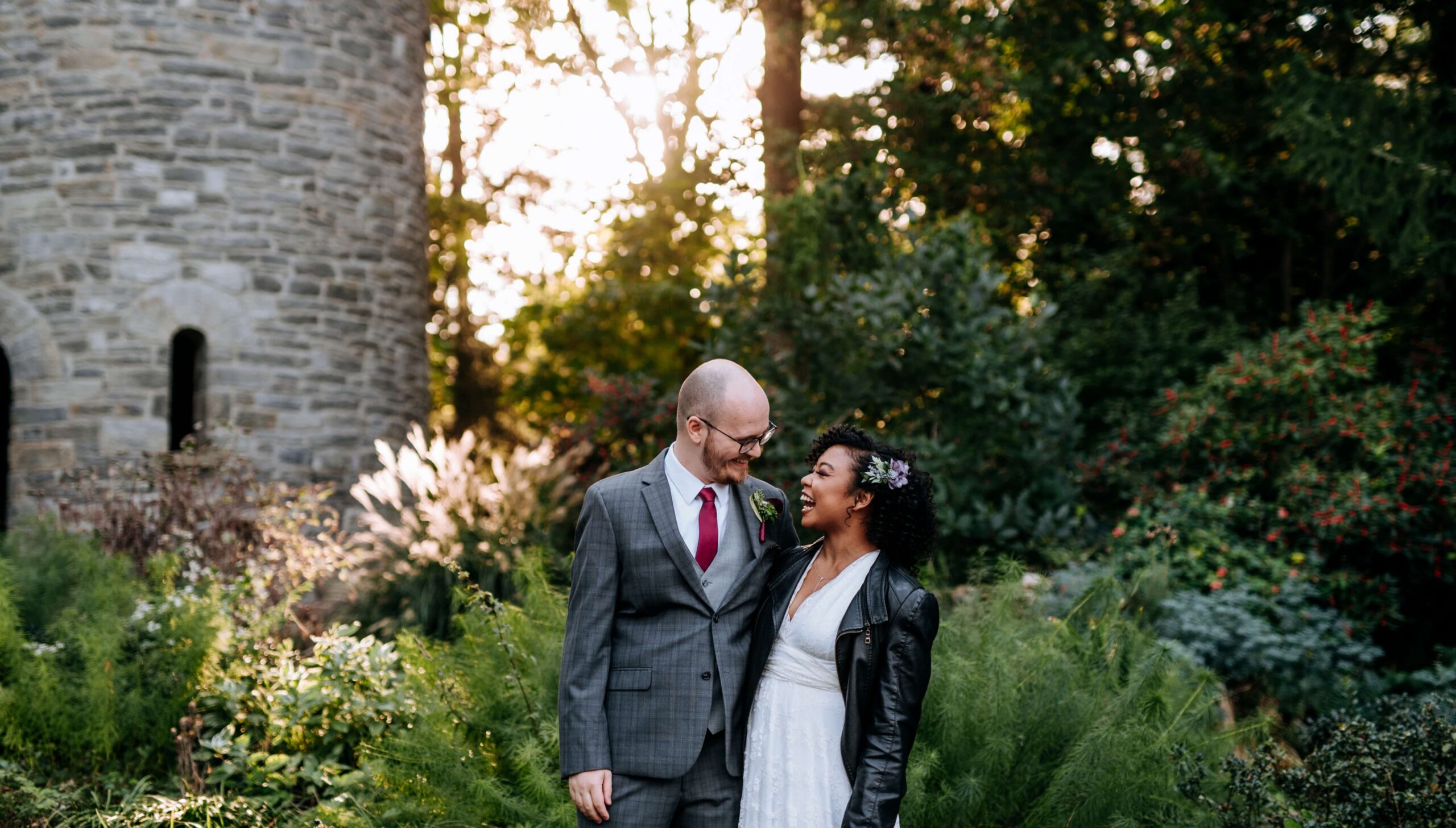 A couple stands smiling at each other in a lush garden with a stone building in the background.