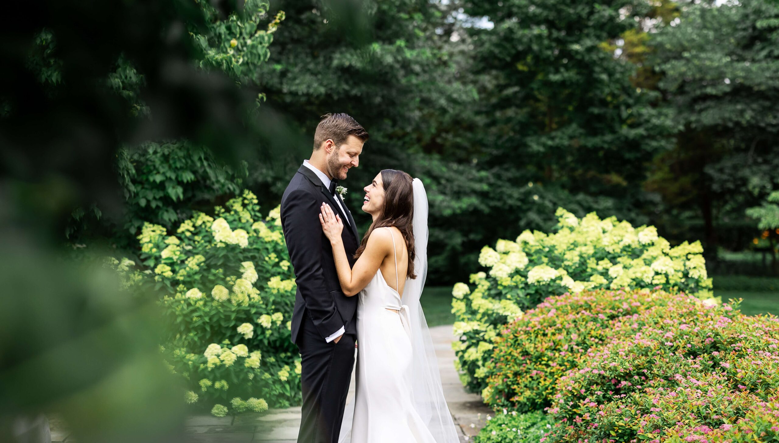 A bride and groom stand in a garden, surrounded by green foliage and flowering bushes. They face each other, smiling, with the groom in a black suit and the bride in a white dress and veil.