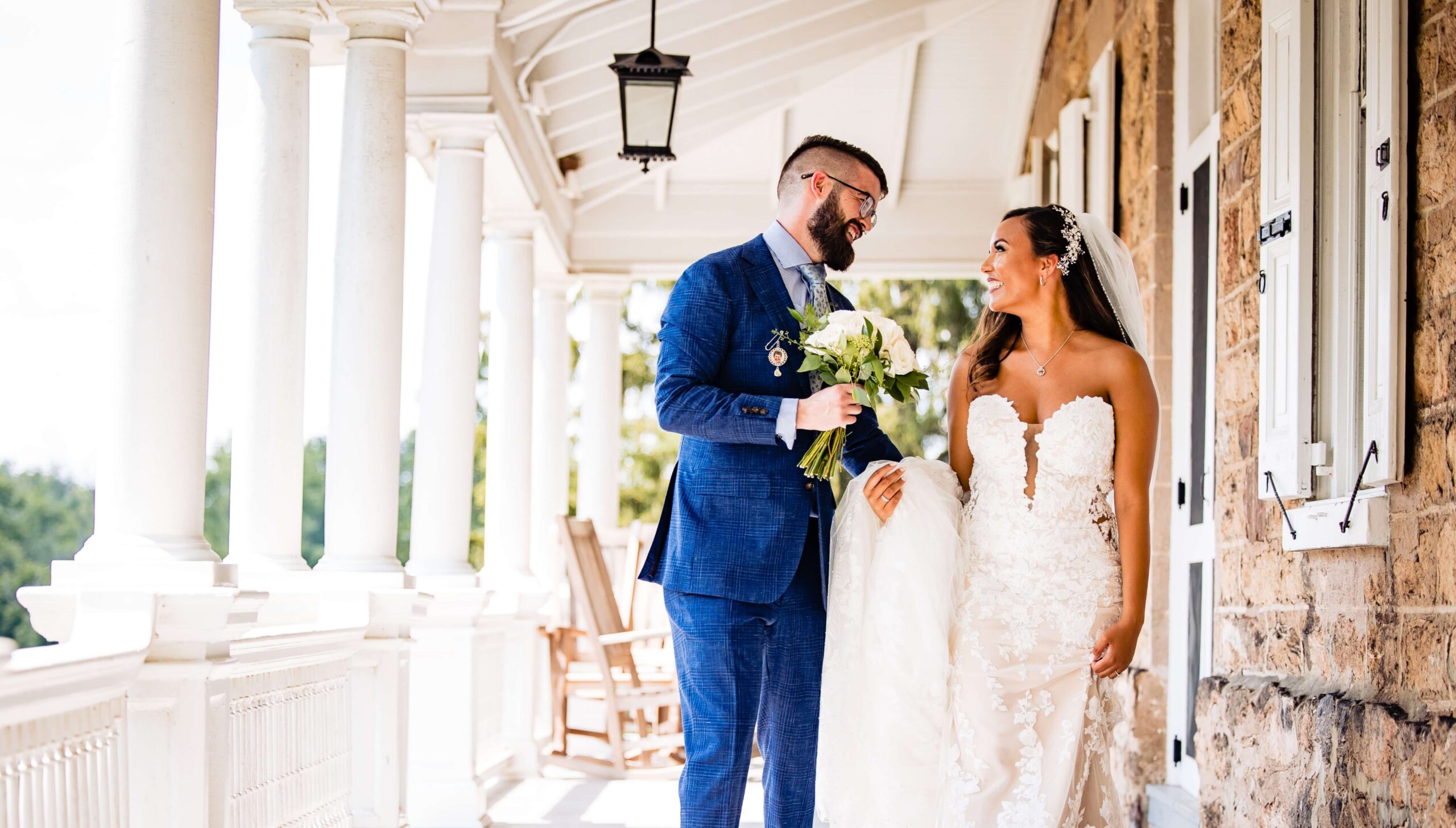 A couple dressed in a suit and wedding gown smile at each other on a porch. The groom holds a bouquet.