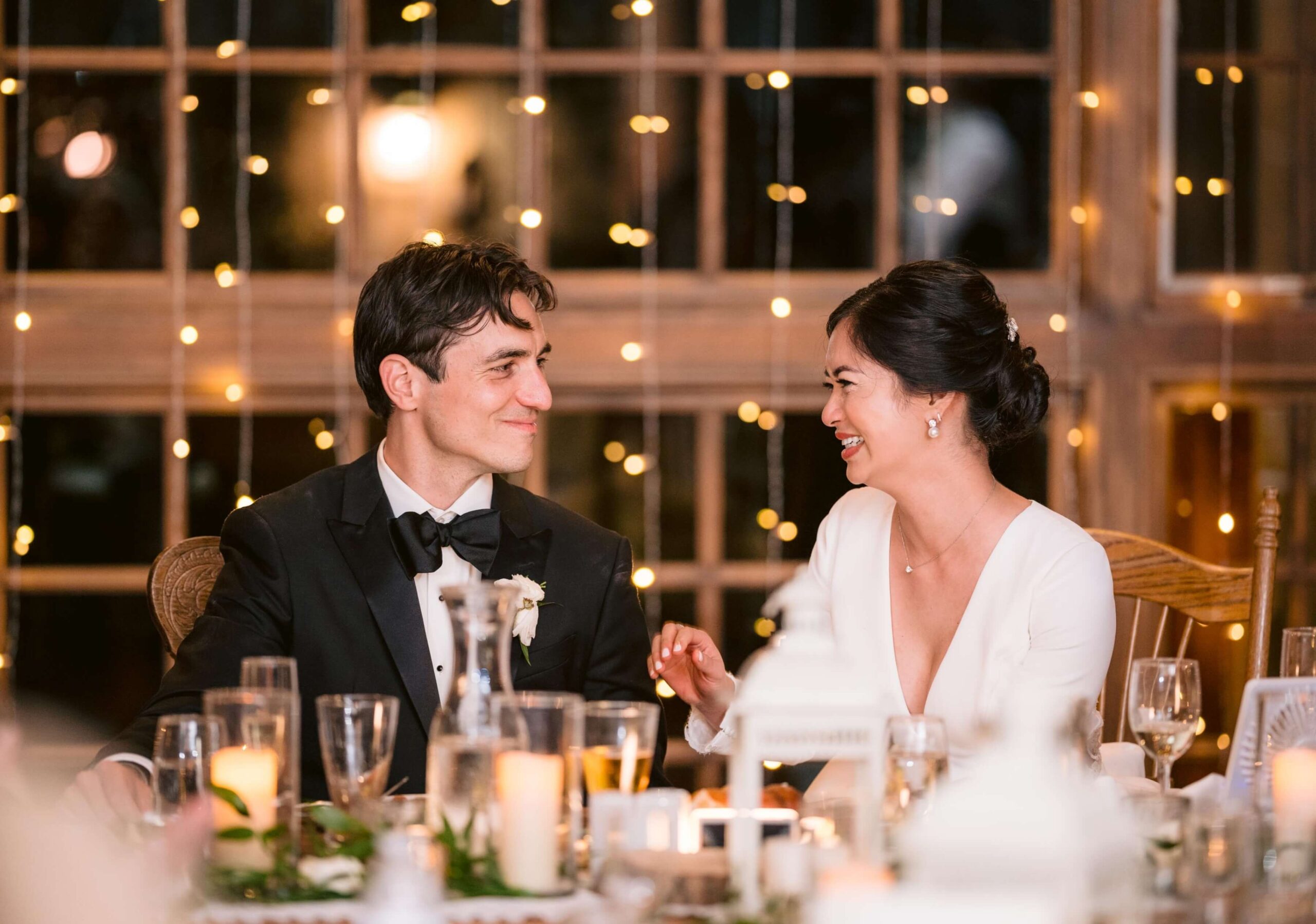 A couple dressed in formal attire sits at a decorated table, smiling at each other. String lights and a wooden window frame are in the background.