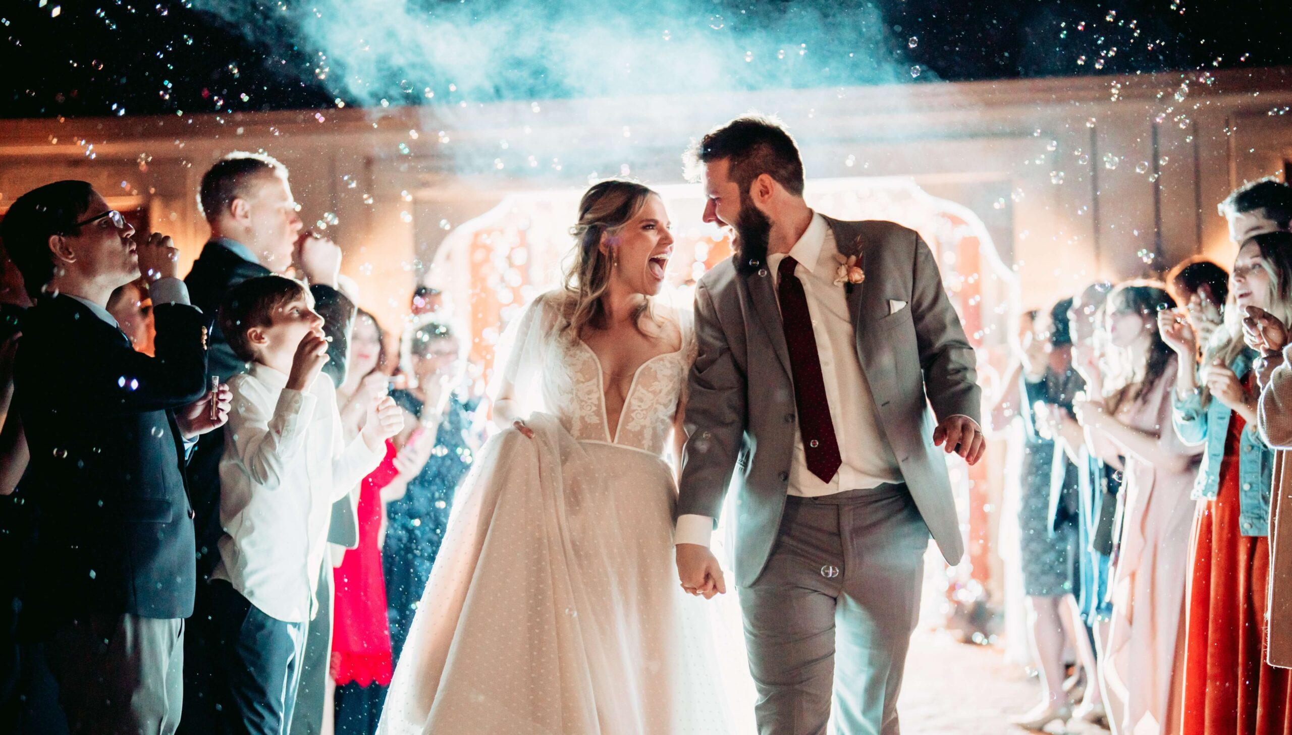 A bride and groom smile and hold hands while walking through a crowd of cheering guests, with bubbles floating around them, in an illuminated festive setting.