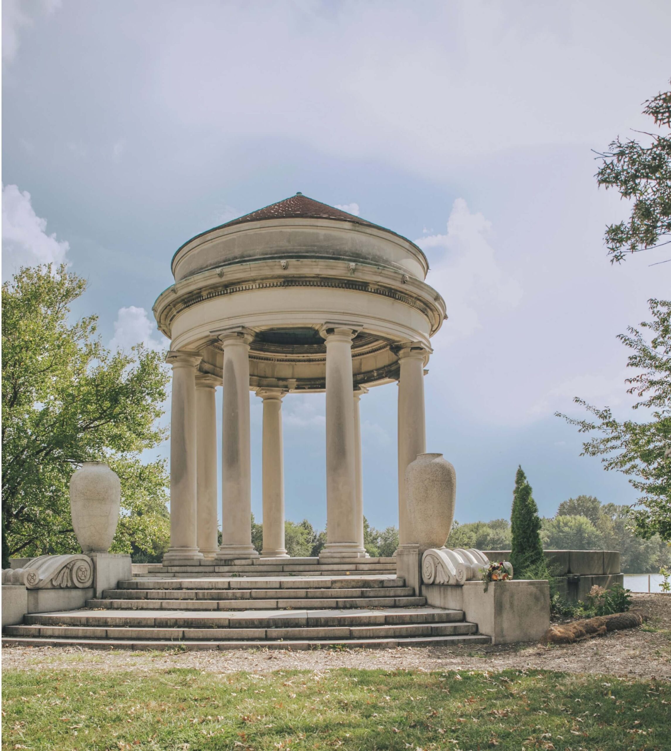 A stone gazebo with columns and a domed roof stands on a grassy area near a body of water, surrounded by trees and large decorative vases.