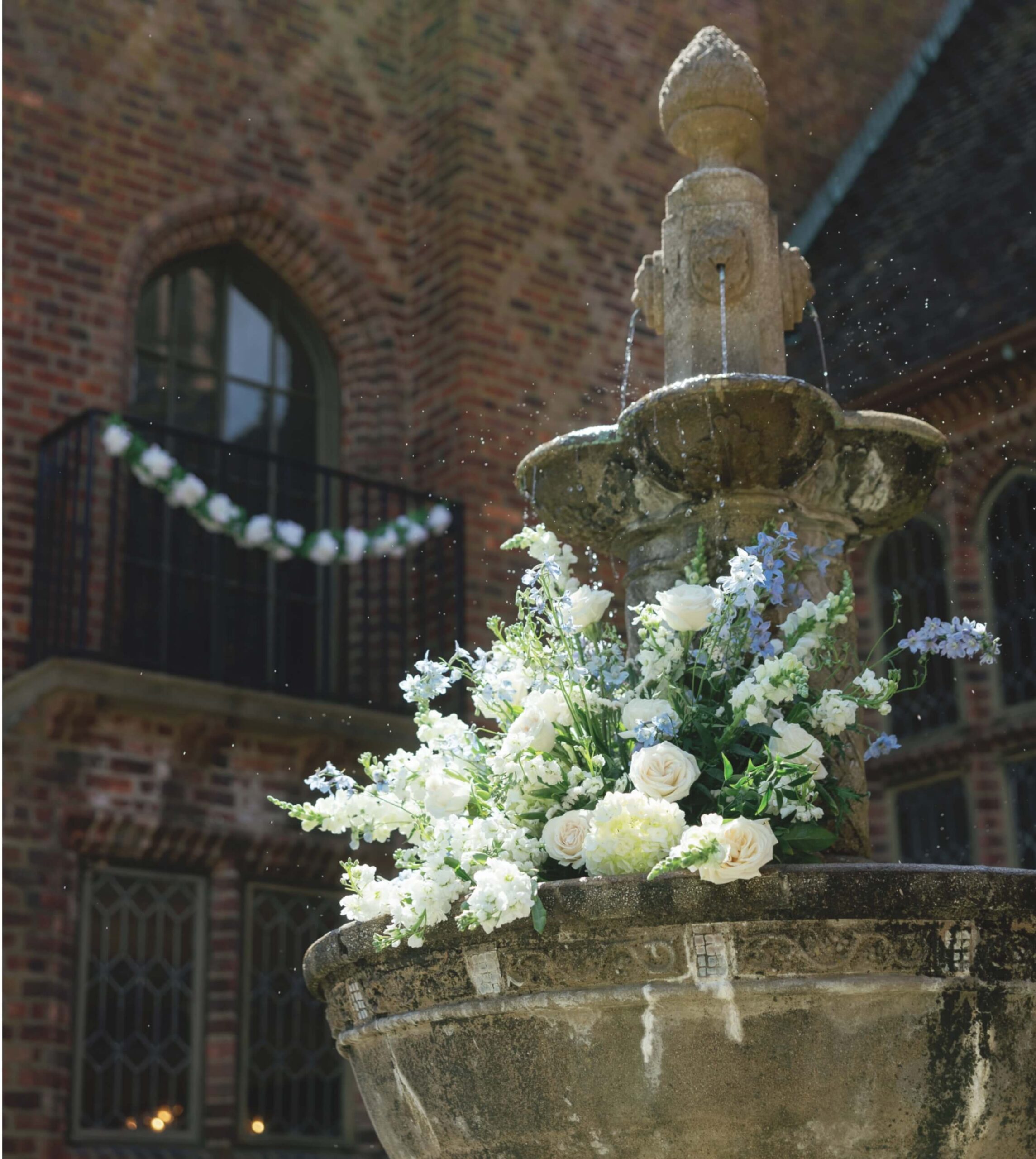 Stone fountain adorned with white and blue flowers in front of a brick building with arched windows and a garland.