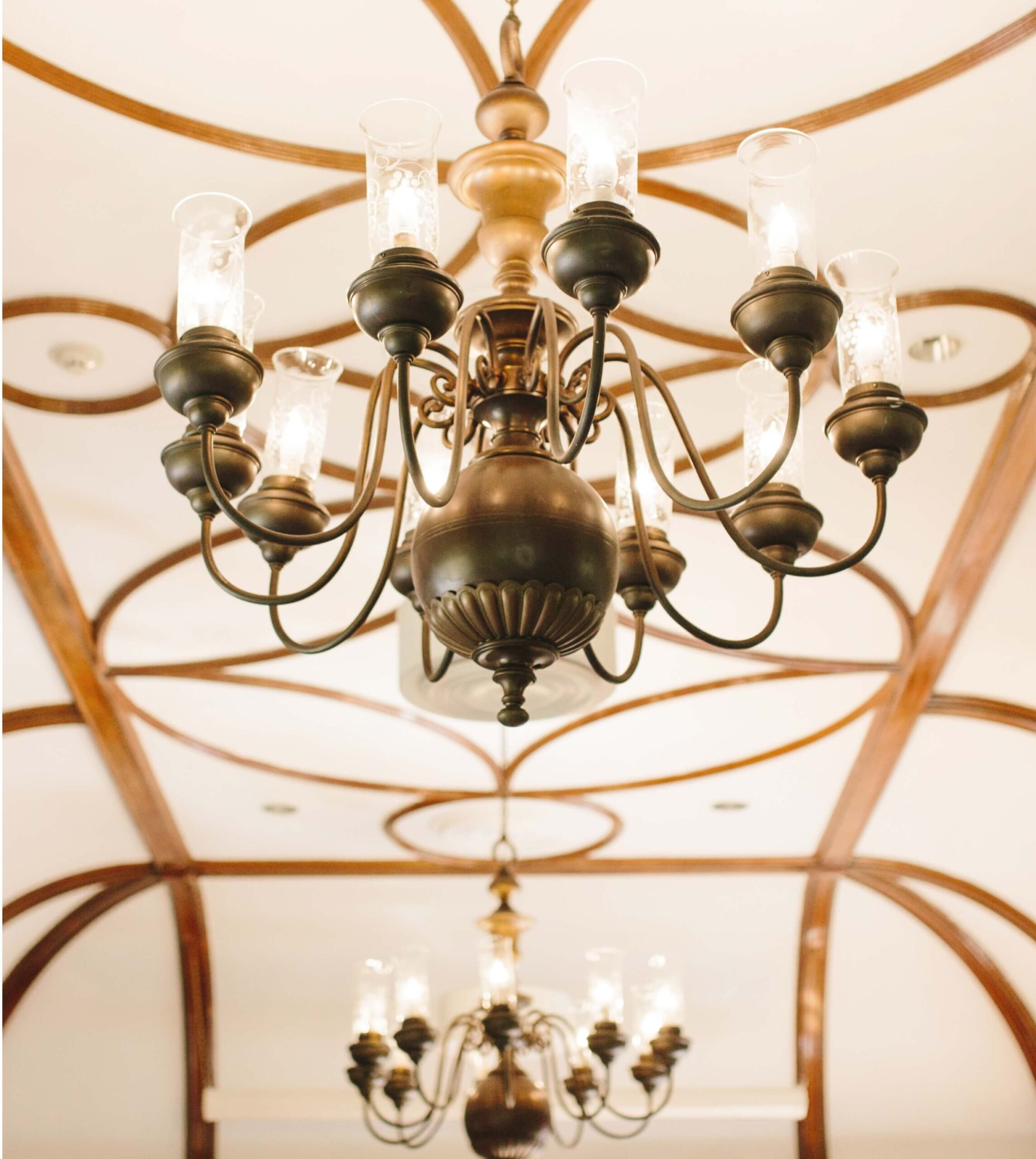 Elegant chandelier with multiple candle-style lights and curved metal arms, suspended from a ceiling with wooden beams in a geometric pattern.