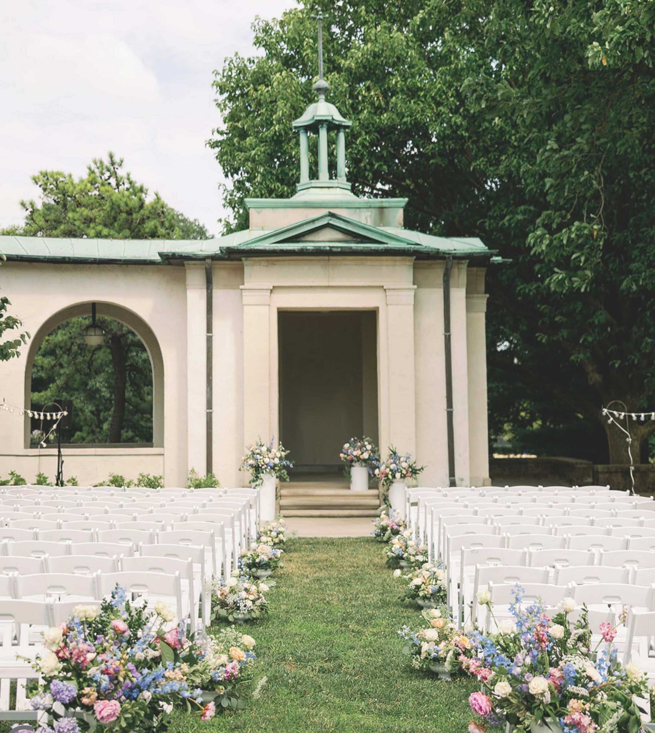 Outdoor wedding ceremony setup with white chairs arranged in rows facing a small gazebo. Aisle lined with colorful floral arrangements. Green trees in the background.