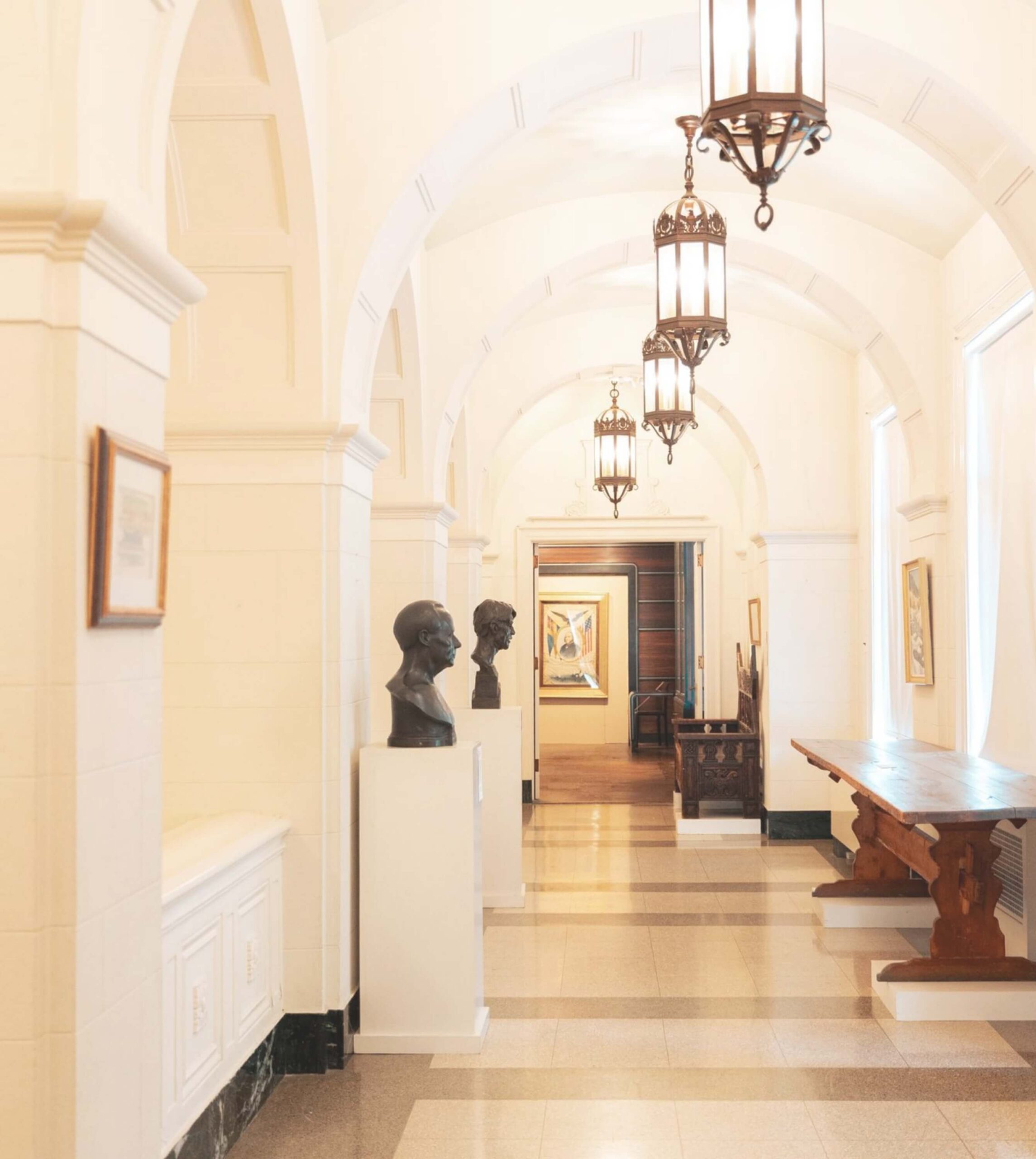A bright, arched hallway features bust sculptures on pedestals, hanging lantern lights, and a wooden table on the right. Paintings adorn the walls, and a doorway is visible at the end.