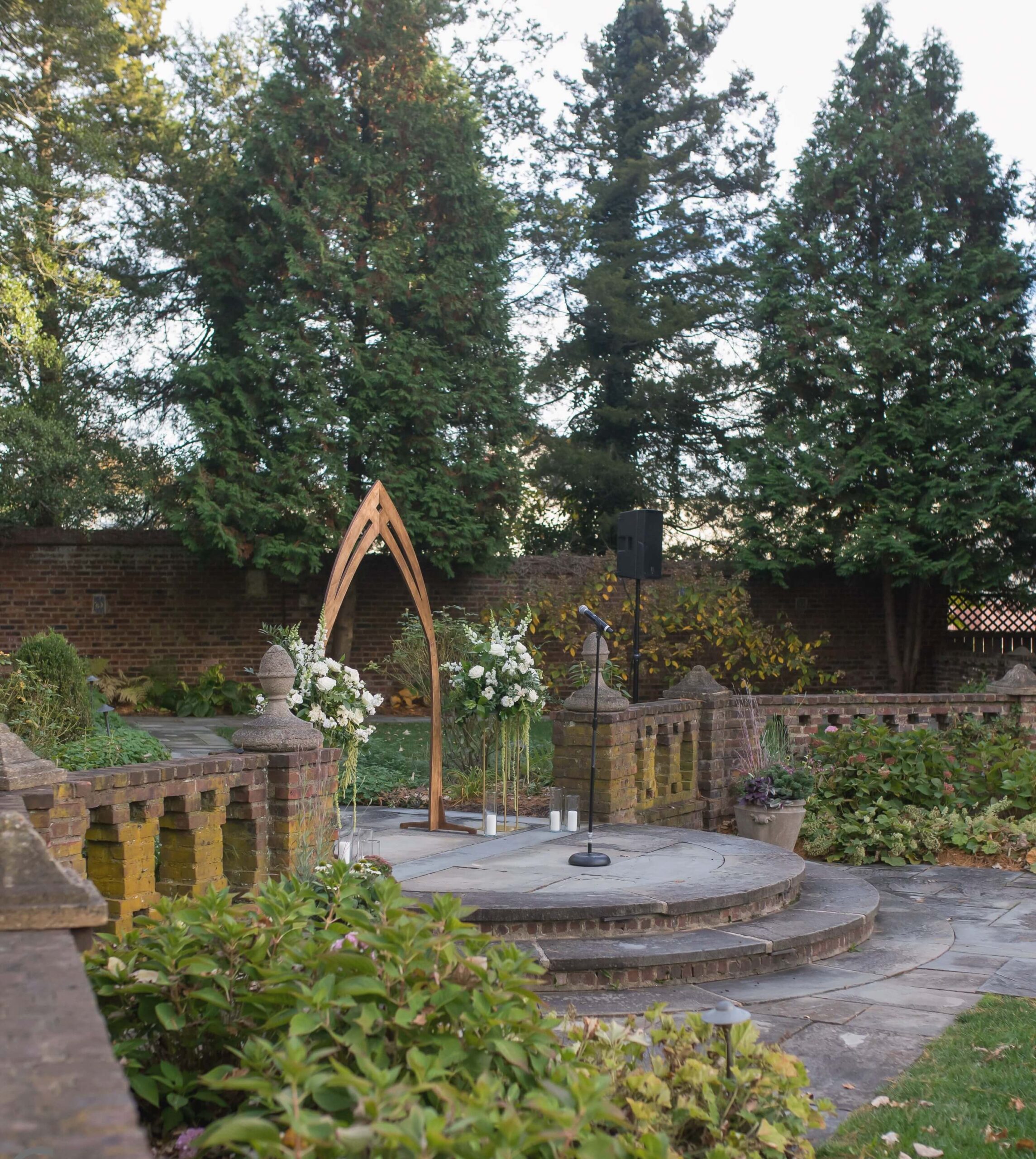 An outdoor wedding ceremony setup with a wooden arch decorated with white flowers on a stone platform, surrounded by greenery and trees.