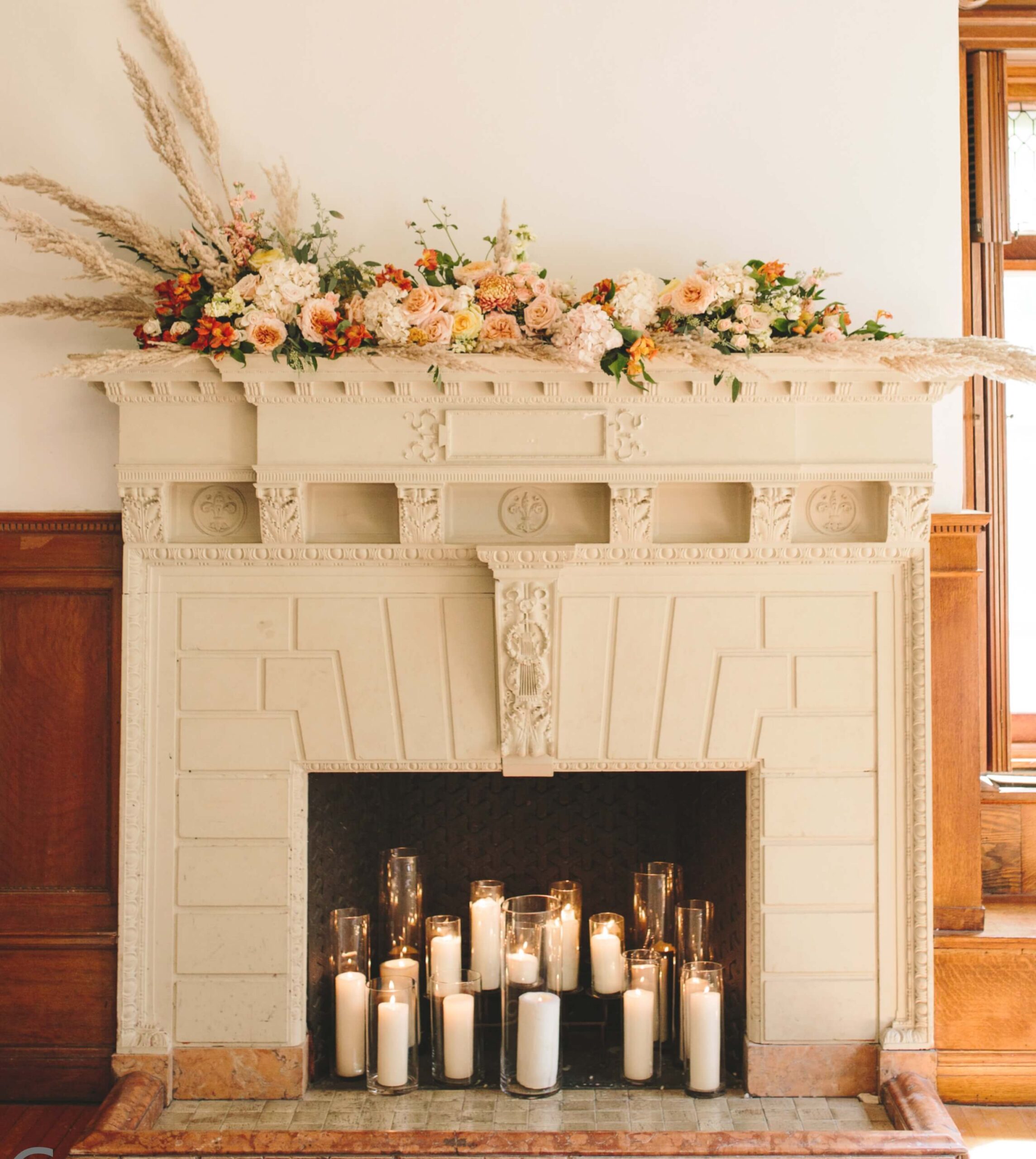 A decorative fireplace with pillar candles inside. The mantel is adorned with a floral arrangement featuring pink and orange flowers and pampas grass.