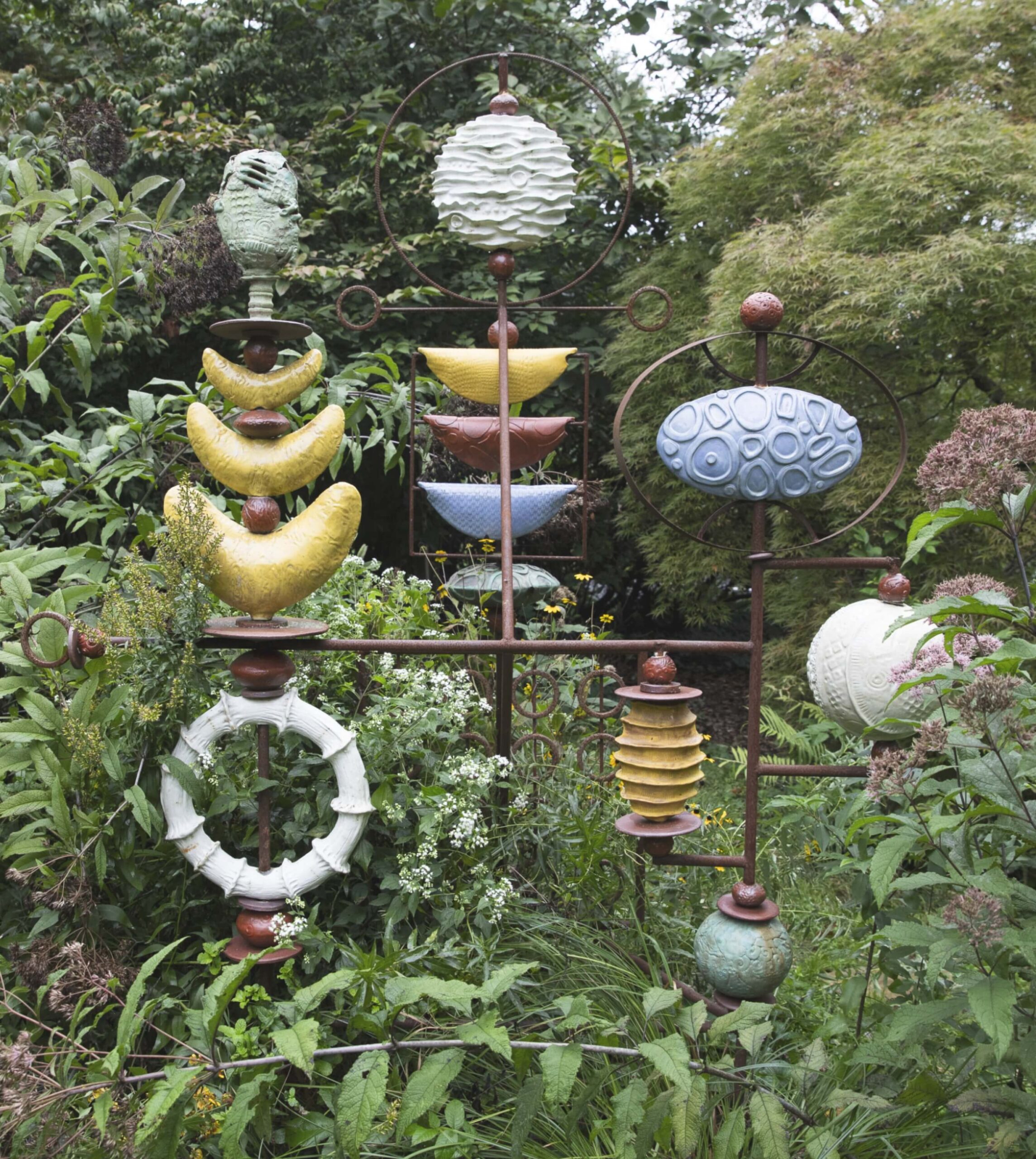 Colorful ceramic garden sculptures with various geometric shapes are displayed on metal stands among lush green foliage.