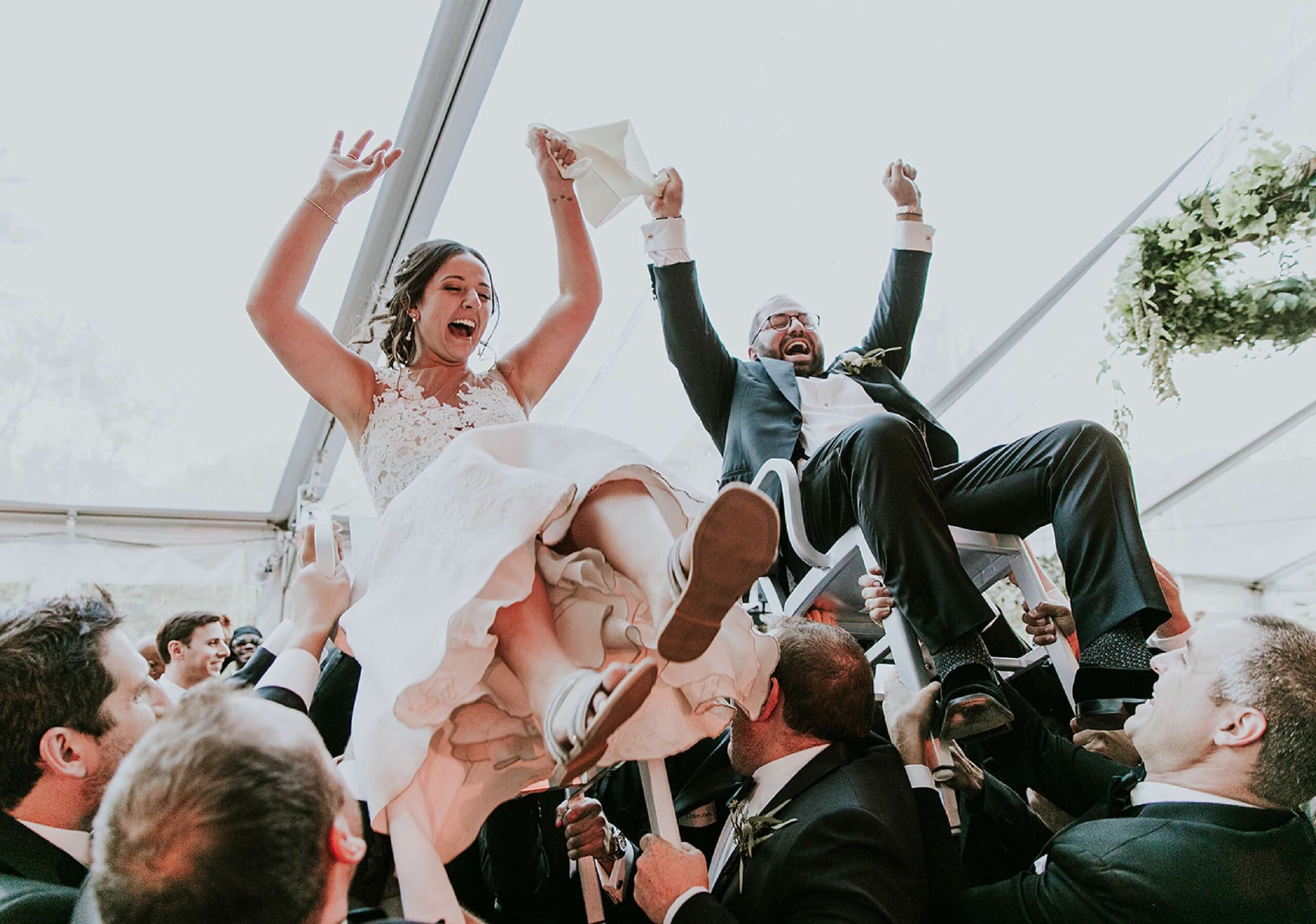 A bride and groom are lifted on chairs by guests during a celebration under a large tent. Both are smiling and raising their arms in joy.