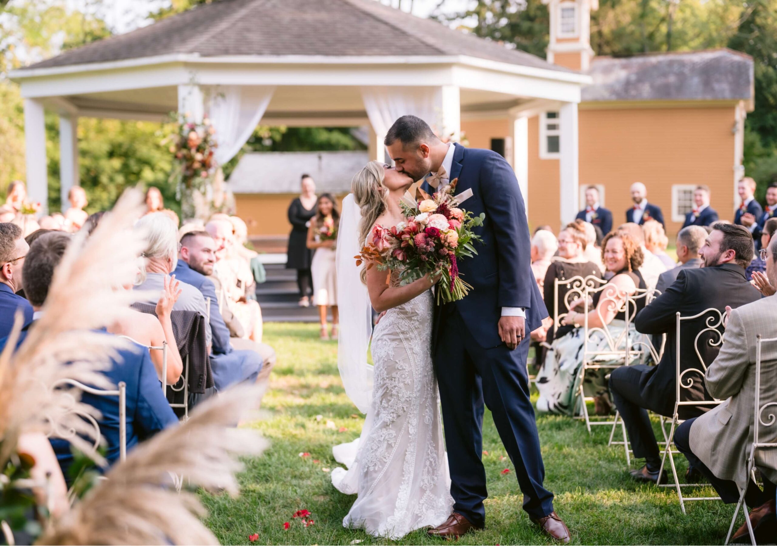 A bride and groom kiss at an outdoor wedding ceremony. Guests seated on chairs watch the couple in front of a gazebo adorned with white drapes and flowers.