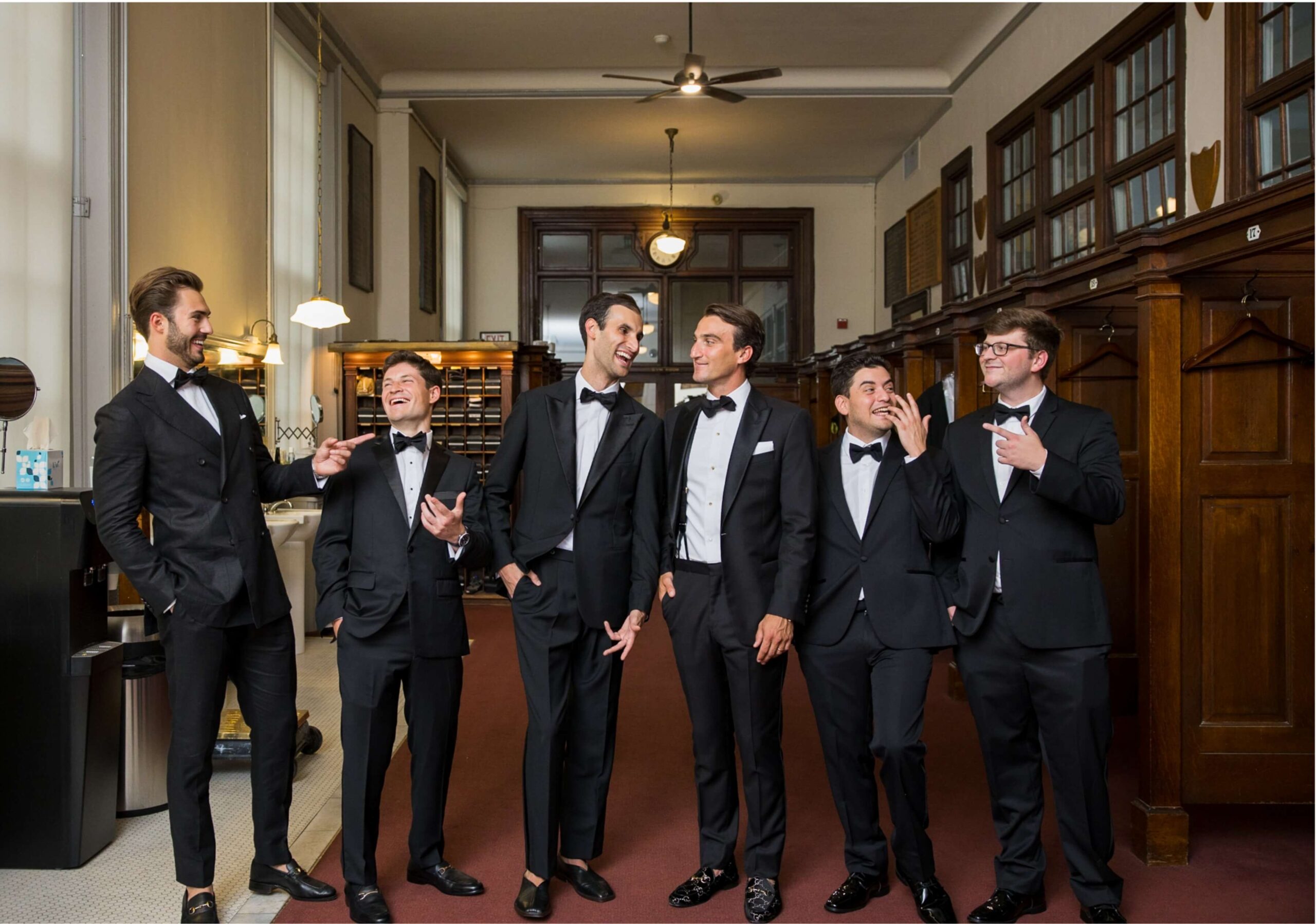 Six men in tuxedos stand and chat in an elegant room with wooden furniture and high ceilings.