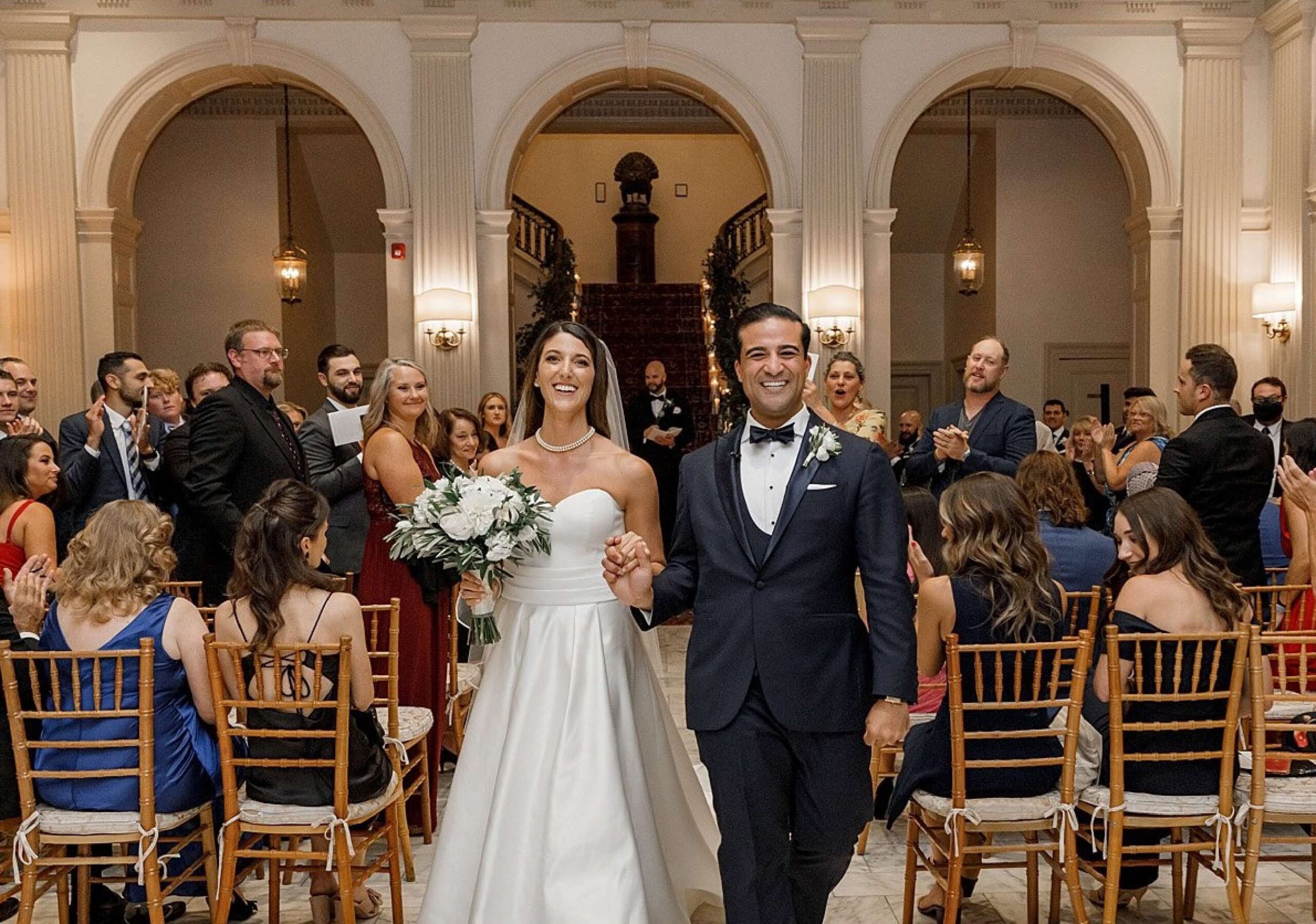 A bride and groom smile while walking down the aisle, holding hands. Guests are seated on either side, clapping in a grand hall with columns and arches.