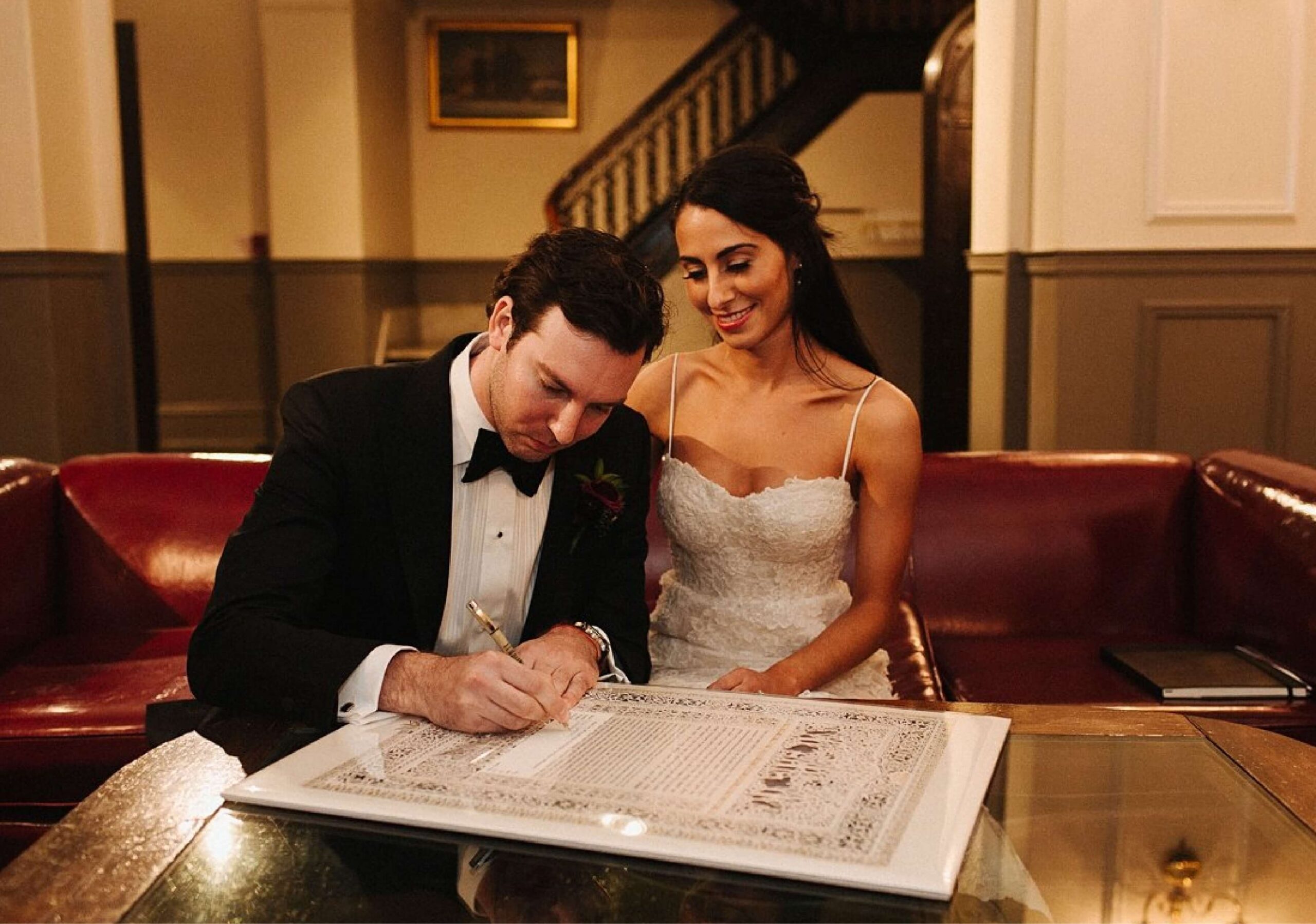 A man in a suit signs a document on a table while a woman in a white dress watches, seated next to him. They are indoors with a staircase visible in the background.
