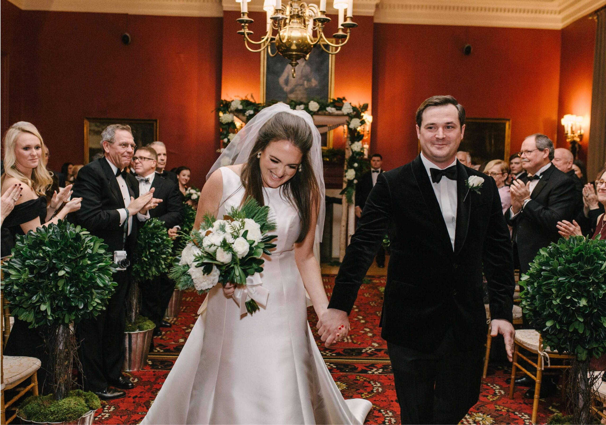 A newlywed couple walks hand in hand down the aisle, smiling. Guests clap and smile in a warmly lit room with red walls and chandeliers.
