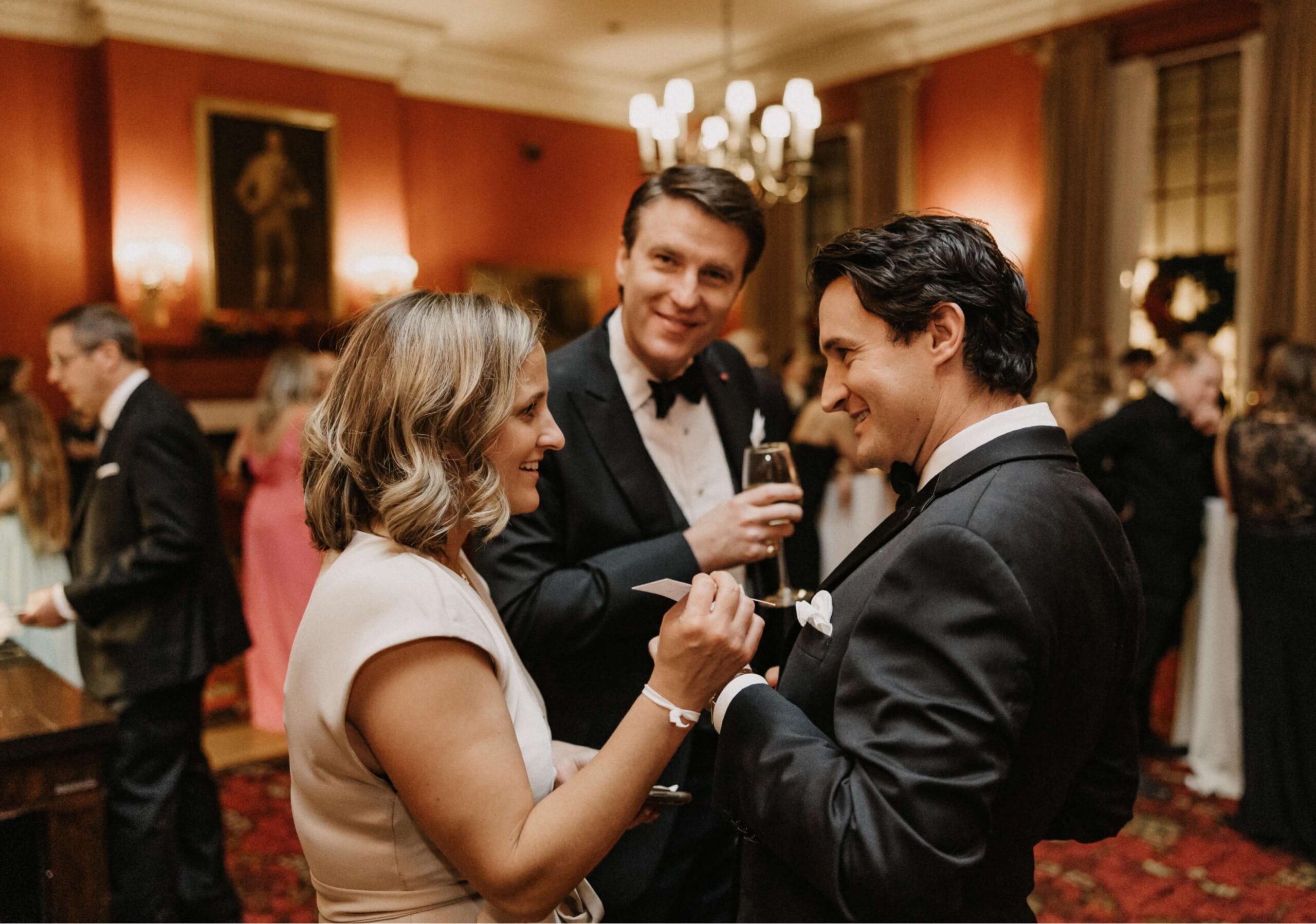 Three people in formal attire converse and smile at a social event in a warmly lit room with a red carpet and chandeliers.
