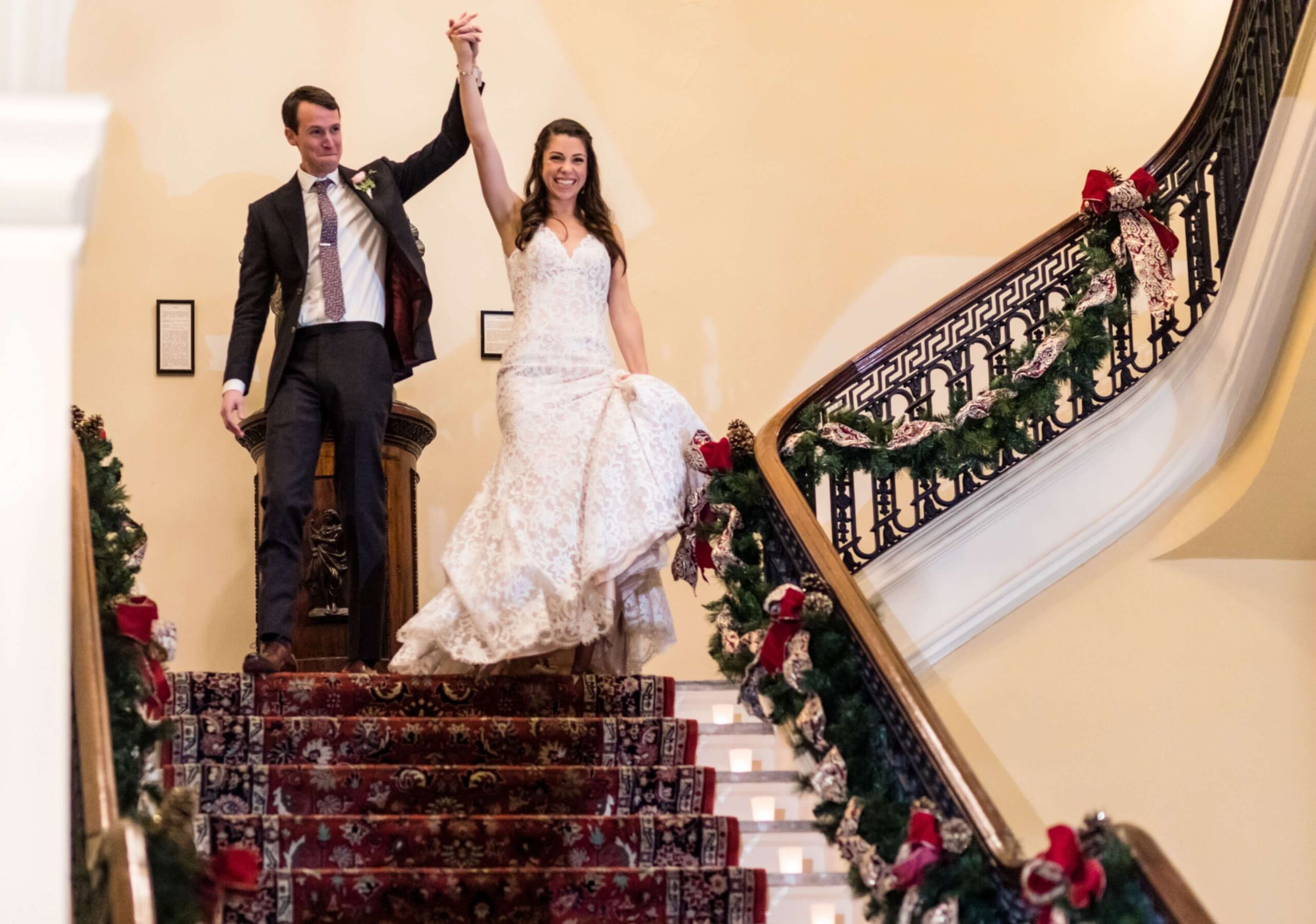 A bride and groom joyfully descend a decorated staircase, hand in hand, with the groom raising their clasped hands in celebration.