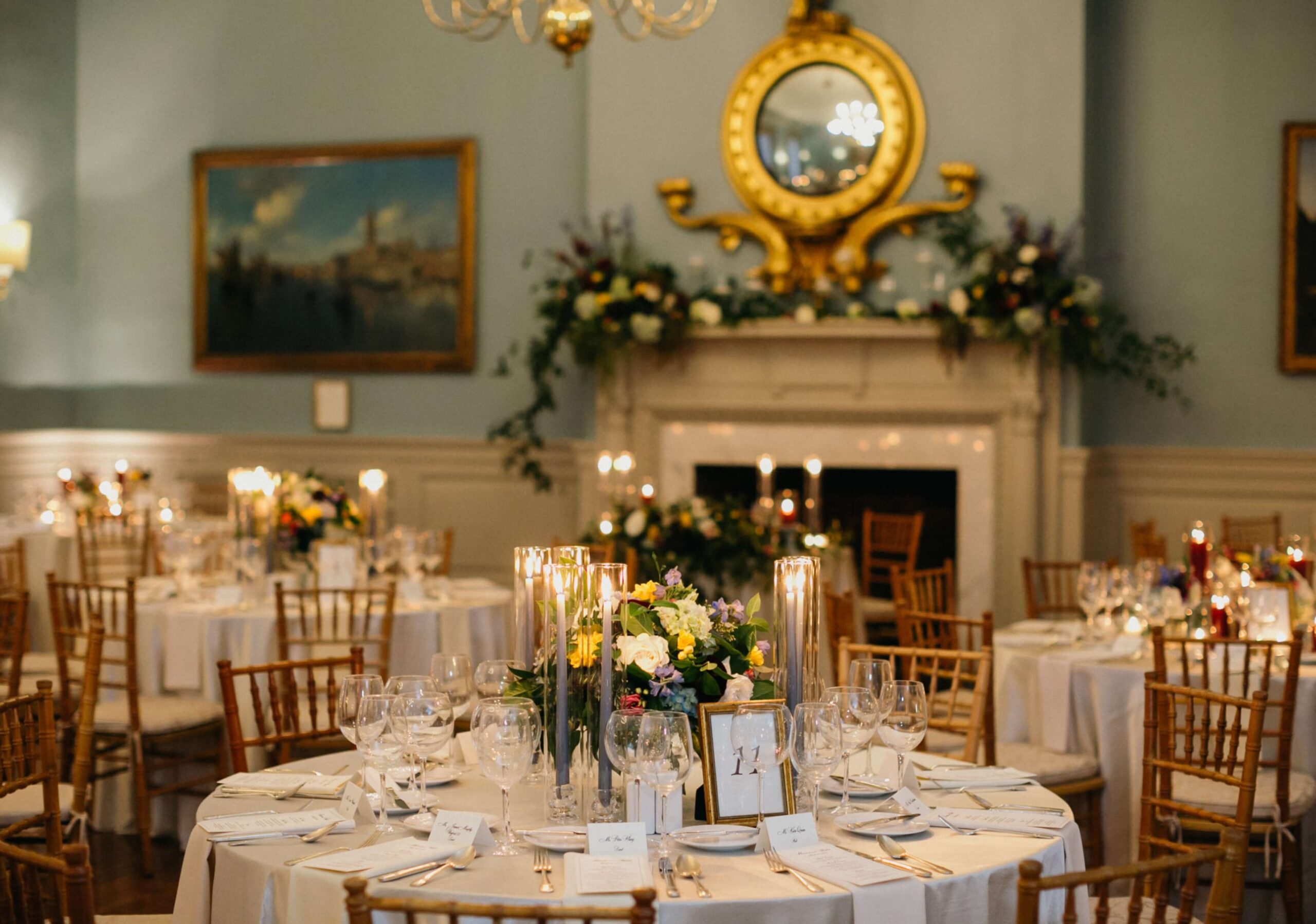 Elegant dining room set for an event with round tables, white tablecloths, floral centerpieces, and wooden chairs. A decorated fireplace and gold mirror are in the background.