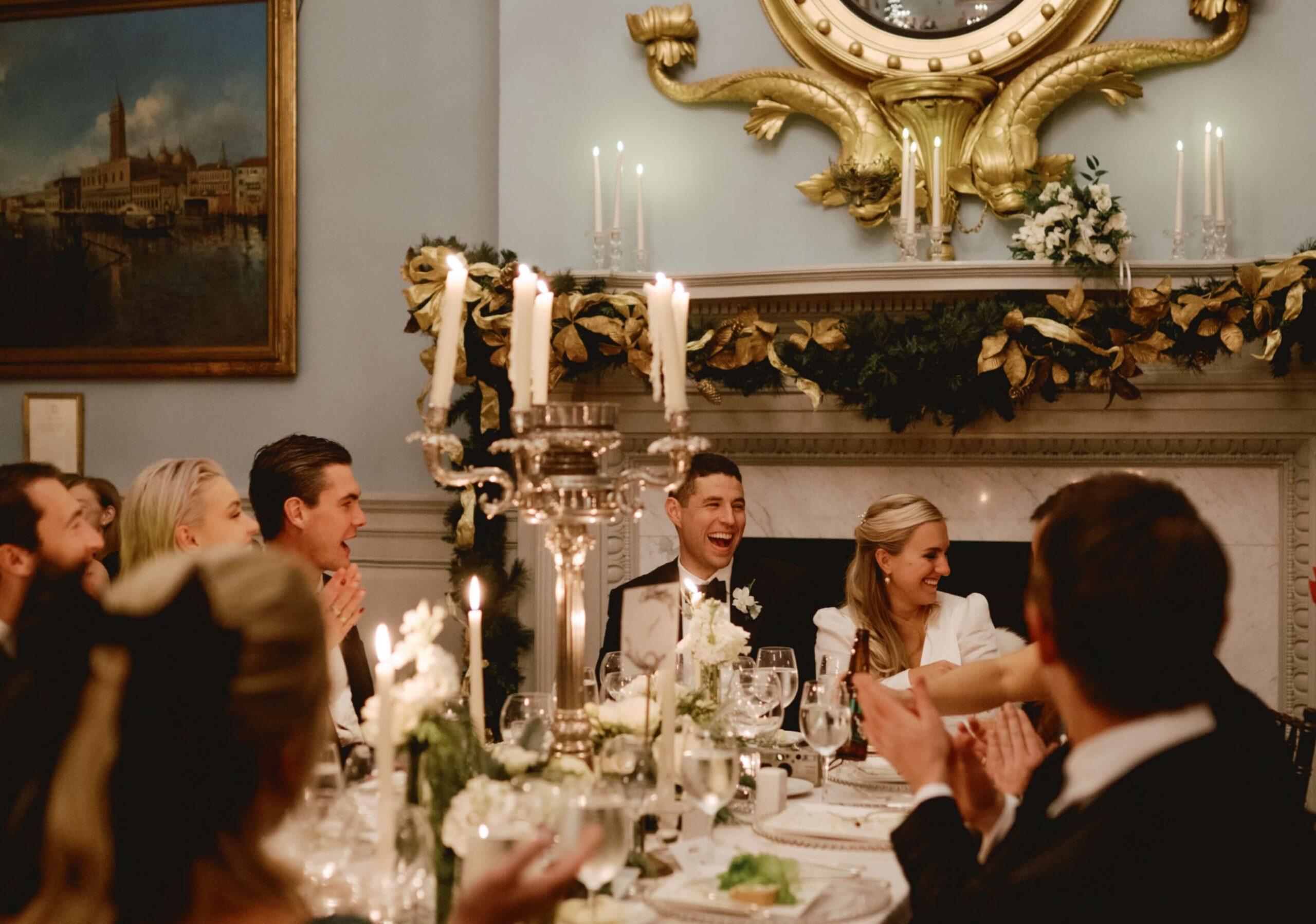 A group of people in formal attire sit around a dining table, clapping and laughing, with candles and elegant decorations in the background.