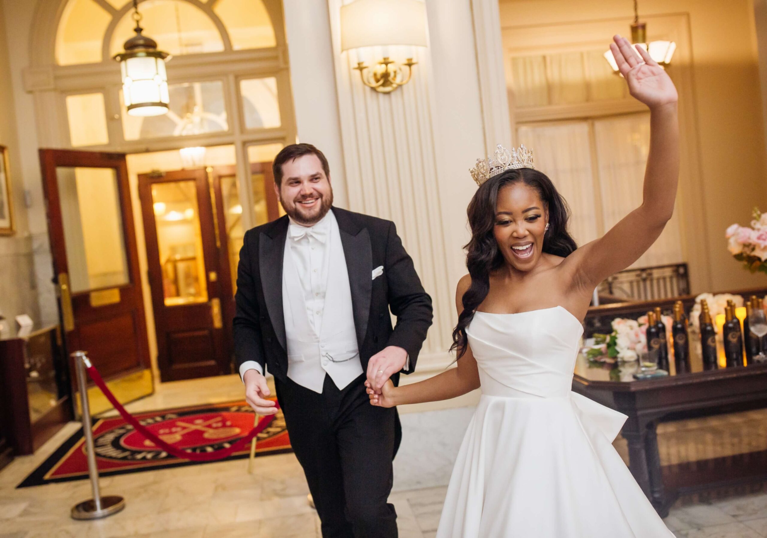 A couple in formal attire, with the woman wearing a tiara and wedding dress, and the man in a tuxedo, joyfully walking indoors.