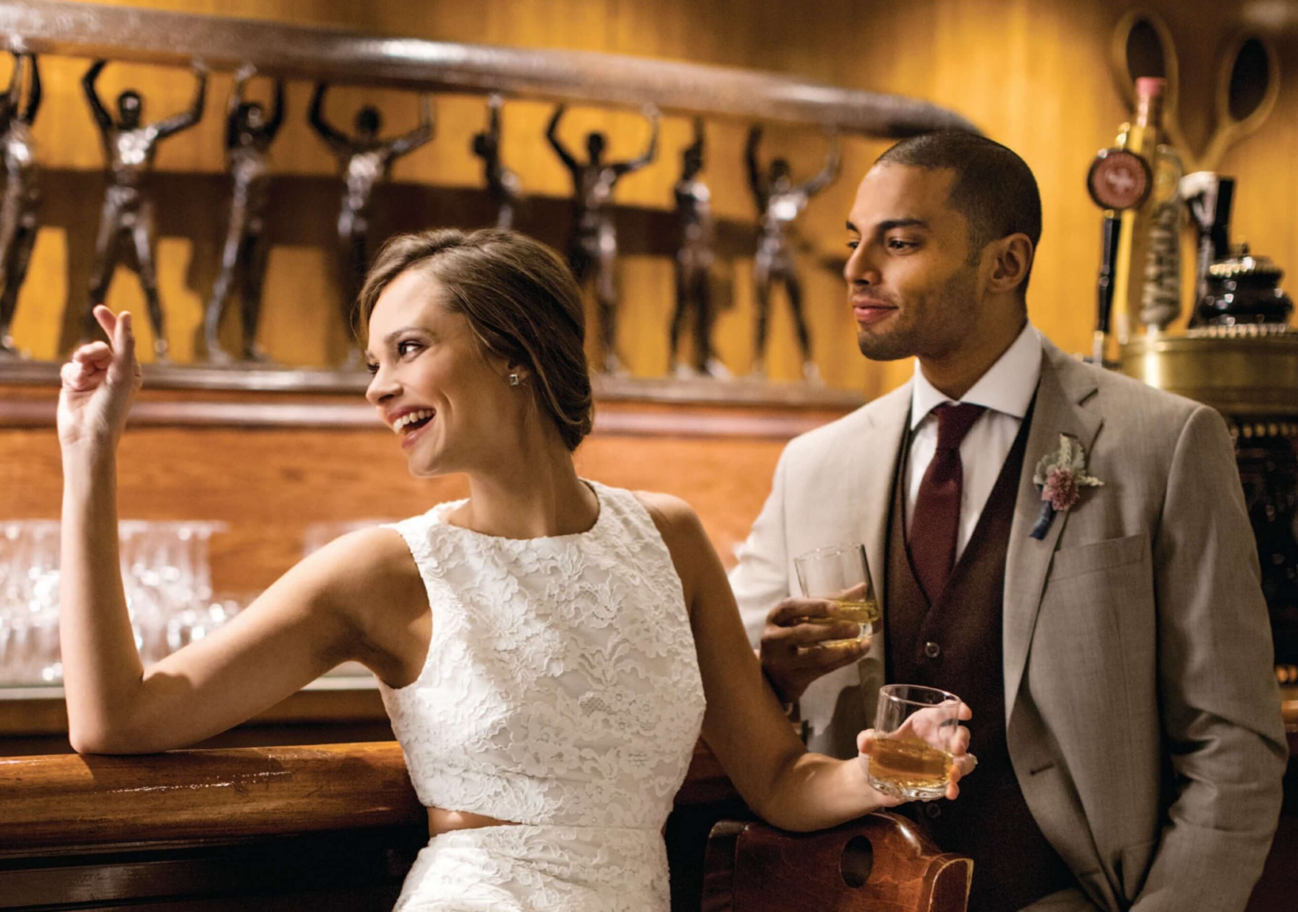 A smiling woman in a white dress gestures at a bar, while a man in a suit stands beside her holding a drink. Bronze sculptures are visible in the background.
