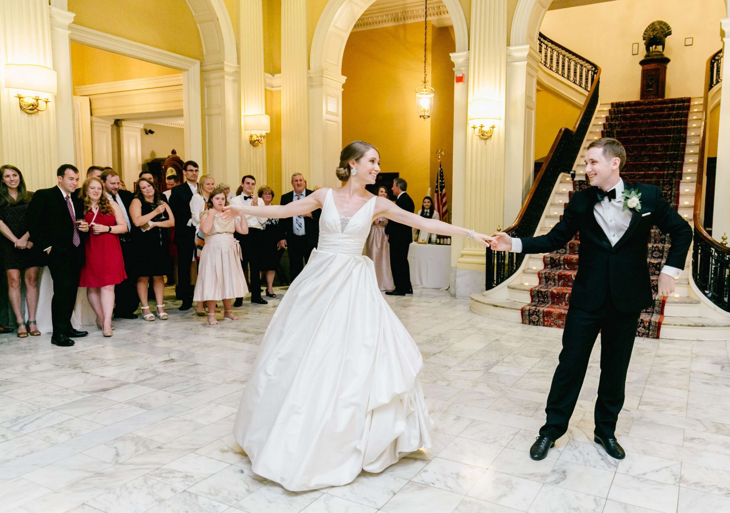 A bride and groom dance in a grand hall with marble floors, surrounded by wedding guests watching and clapping.