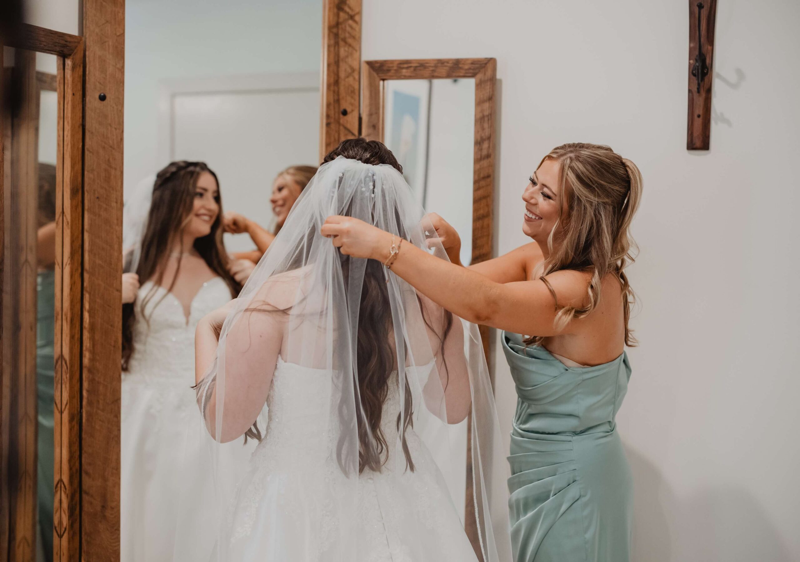 A bridesmaid in a green dress adjusts the veil of a smiling bride in a white gown standing in front of a mirror.