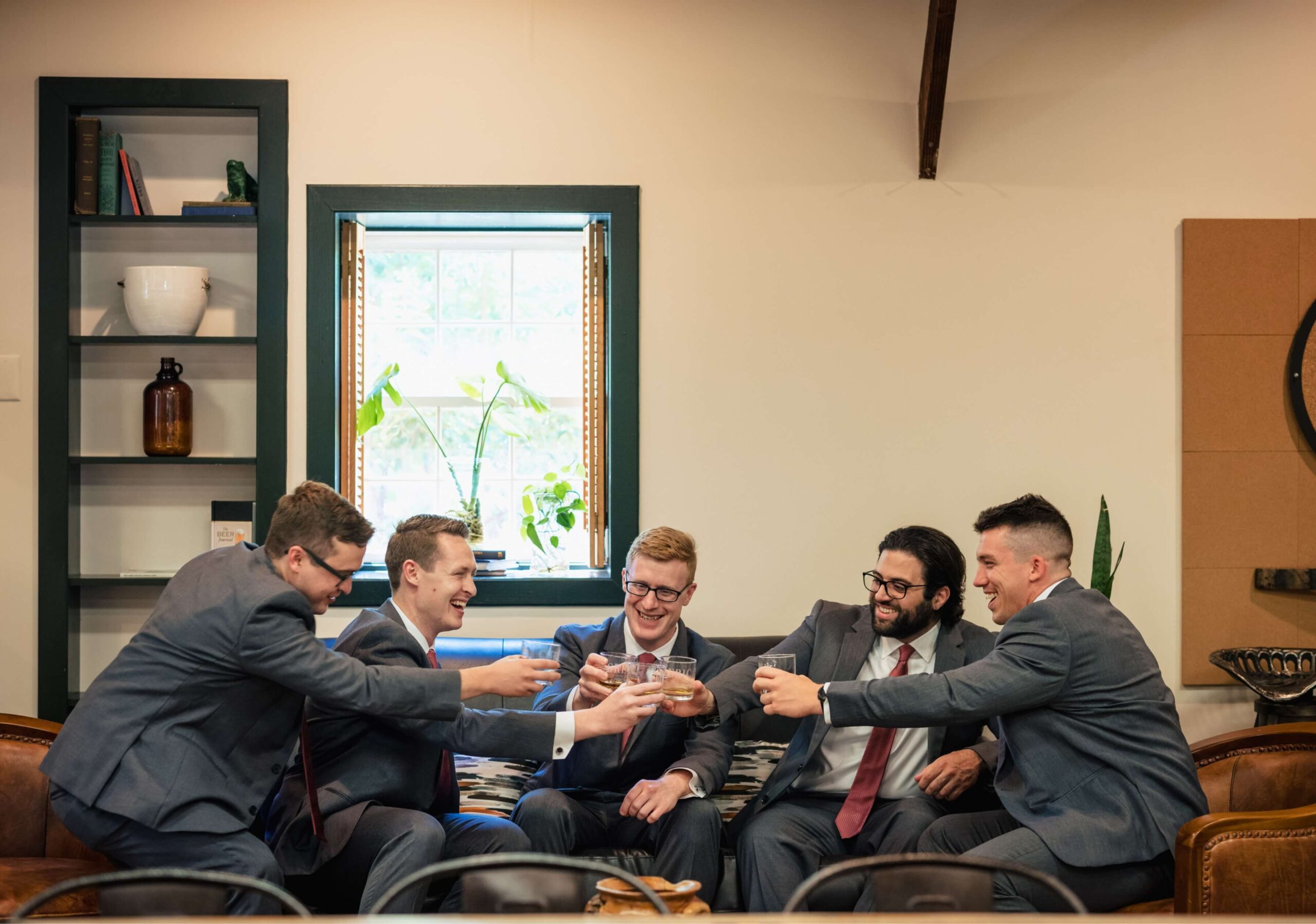 Five men in suits sitting on a couch, smiling and clinking glasses in a well-lit room with shelves and plants.