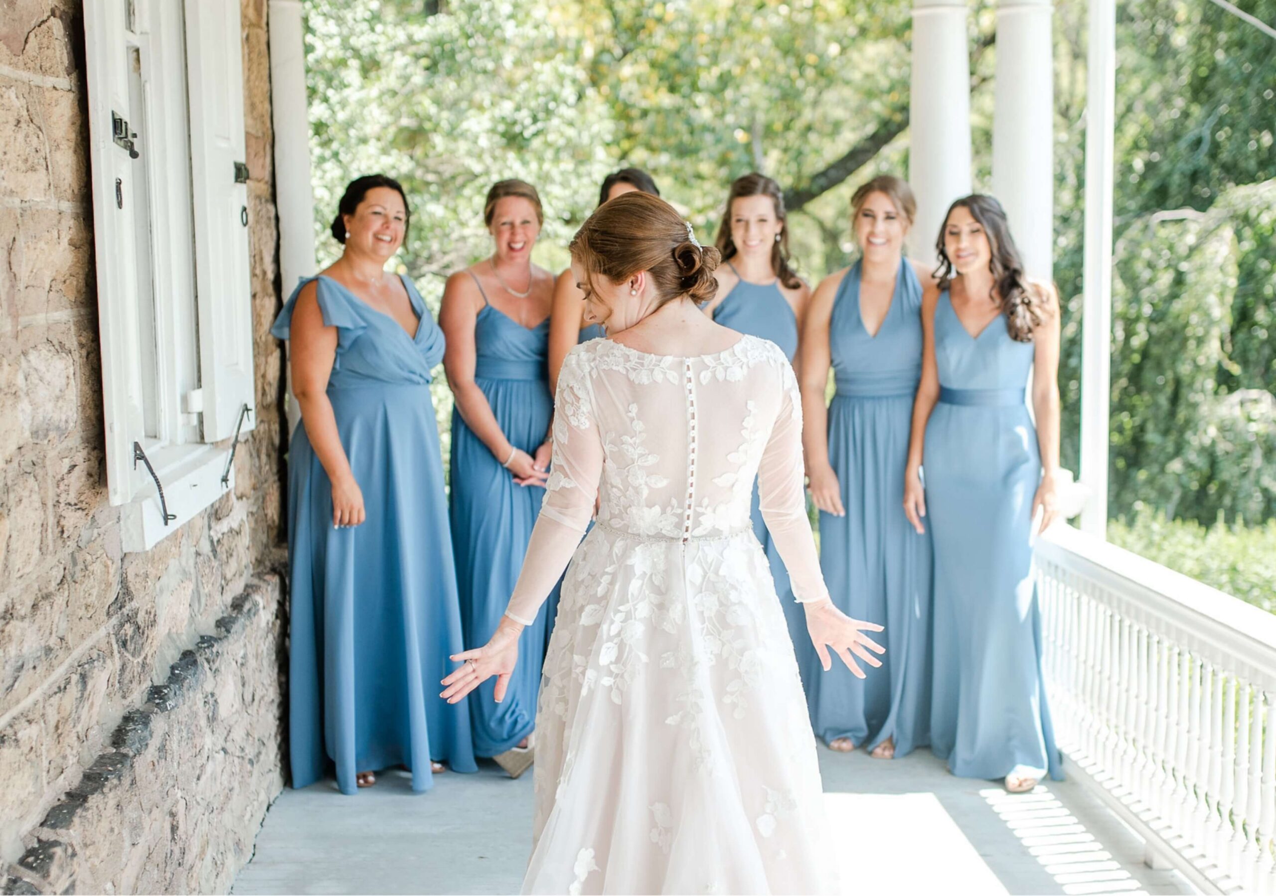 A bride in a white dress faces six bridesmaids in blue dresses on a sunlit porch.