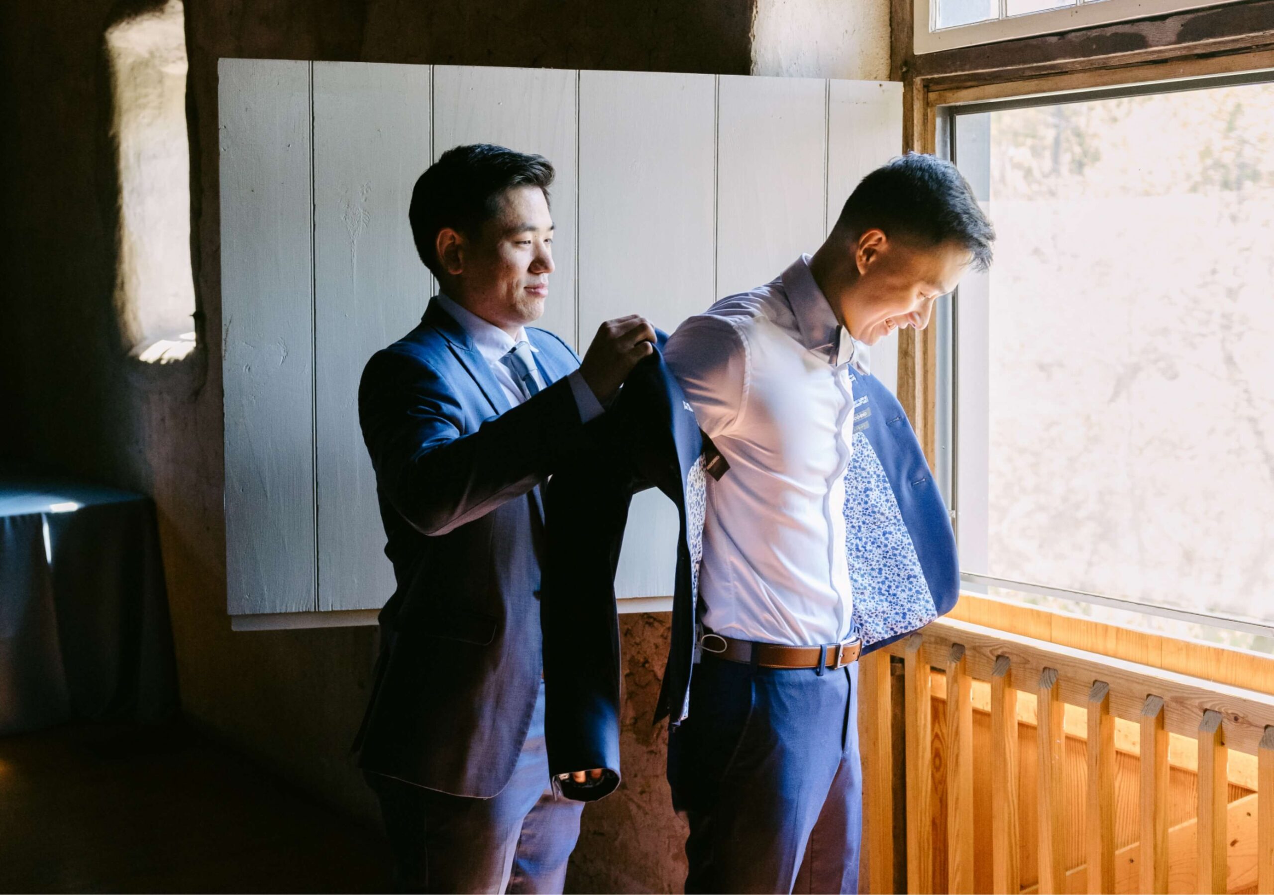 A man in a suit helps another man put on a jacket in a sunlit room by a window.