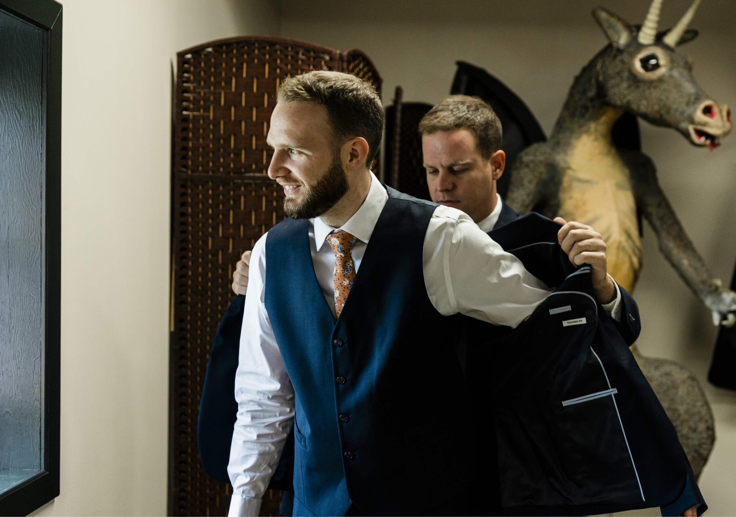 Two men in a room; one adjusts his suit while another helps him. A stuffed donkey is visible in the background.