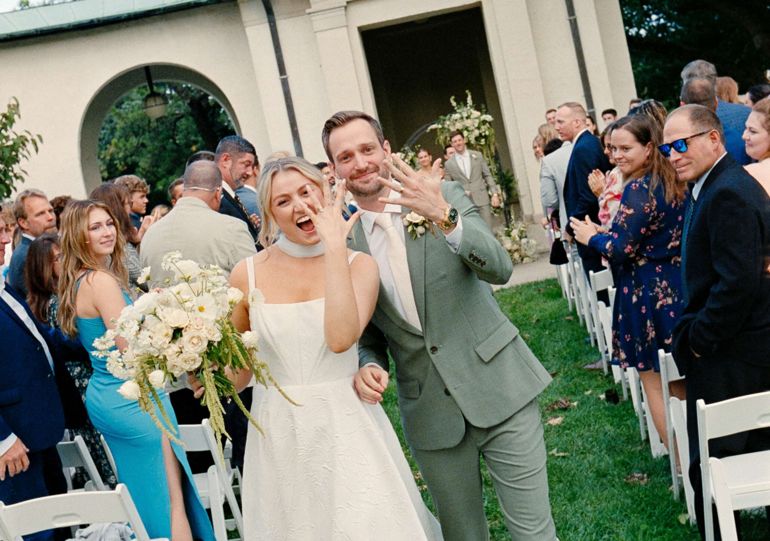 A bride and groom smile and show their rings to the camera while walking down an aisle outdoors, surrounded by guests.